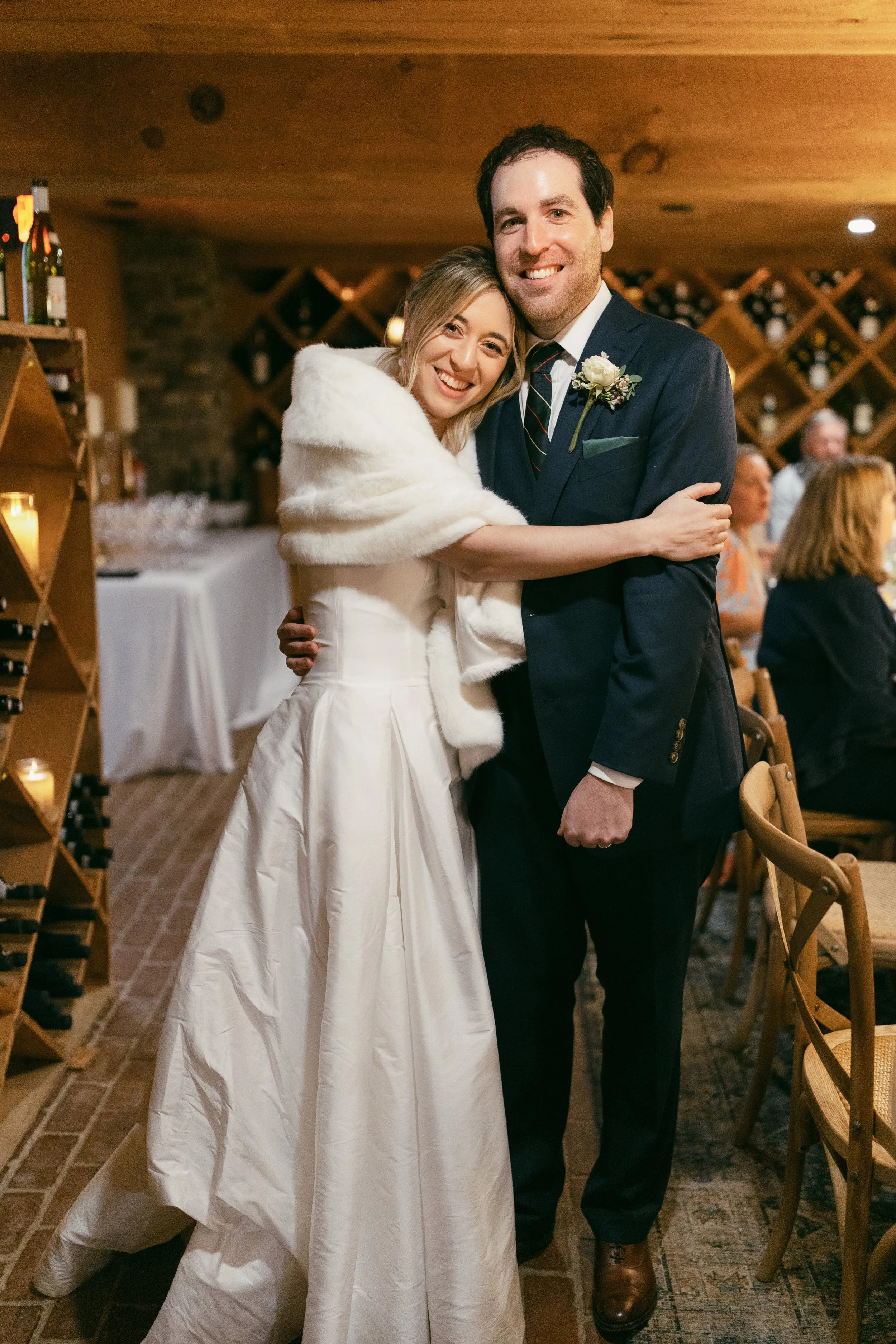 A bride and groom hugging at their wedding reception, surrounded by guests, wooden wine rack, and a decorated indoor venue.