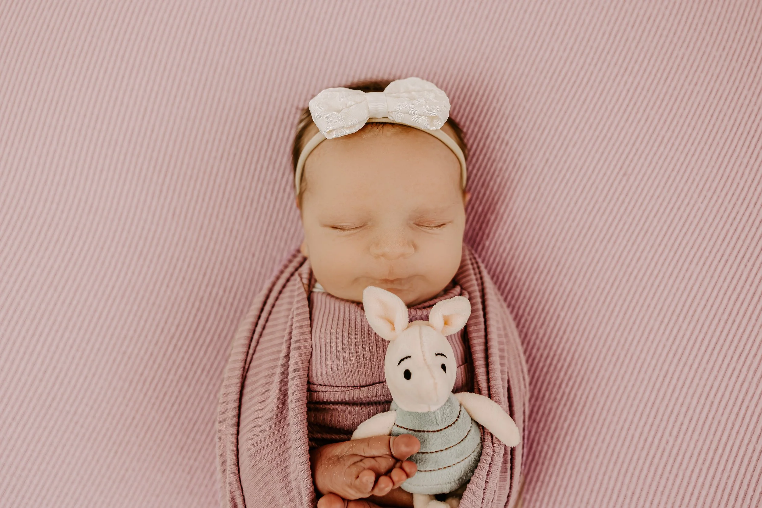 Sleeping newborn baby girl wearing a white headband with a bow, wrapped in a pink blanket, holding a stuffed animal