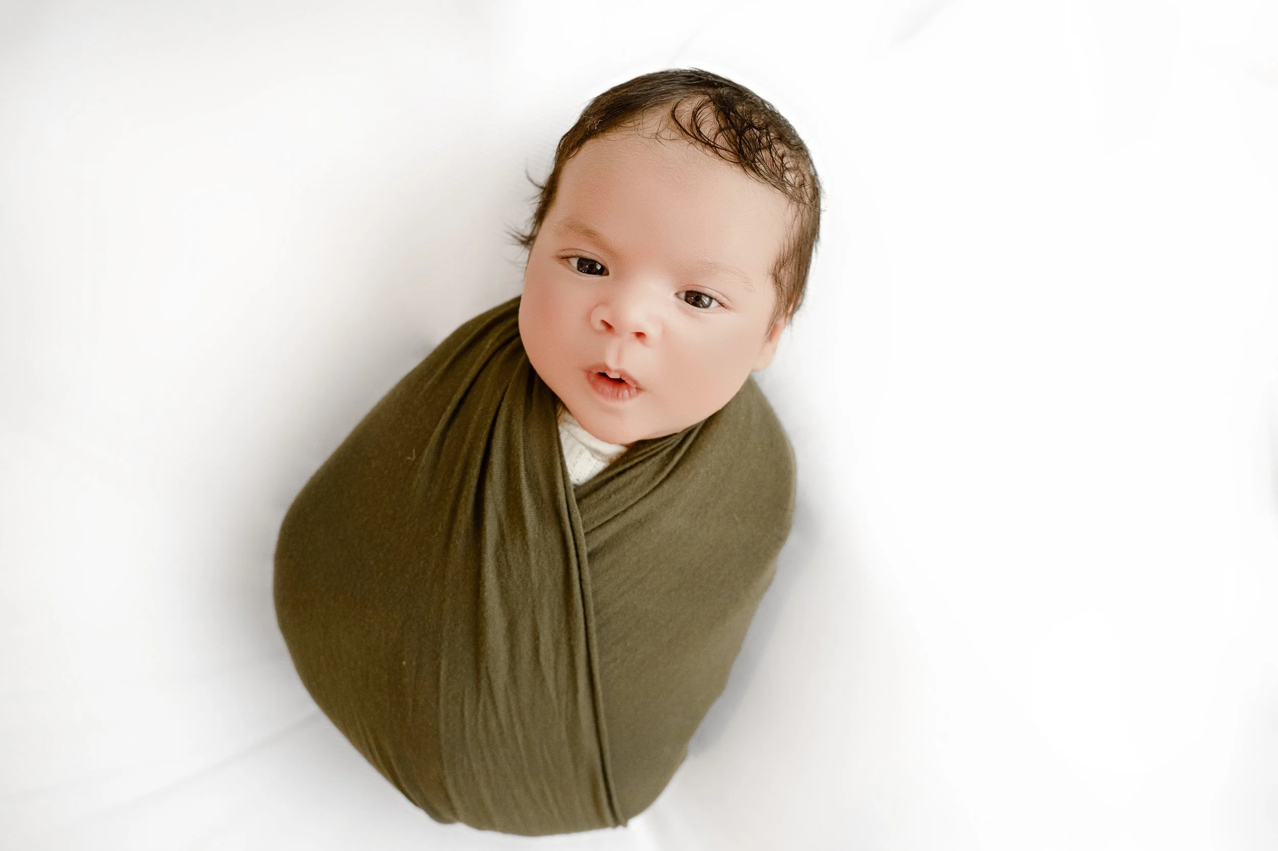 Young baby wrapped in an olive green blanket on a white background, looking up with a surprised expression.