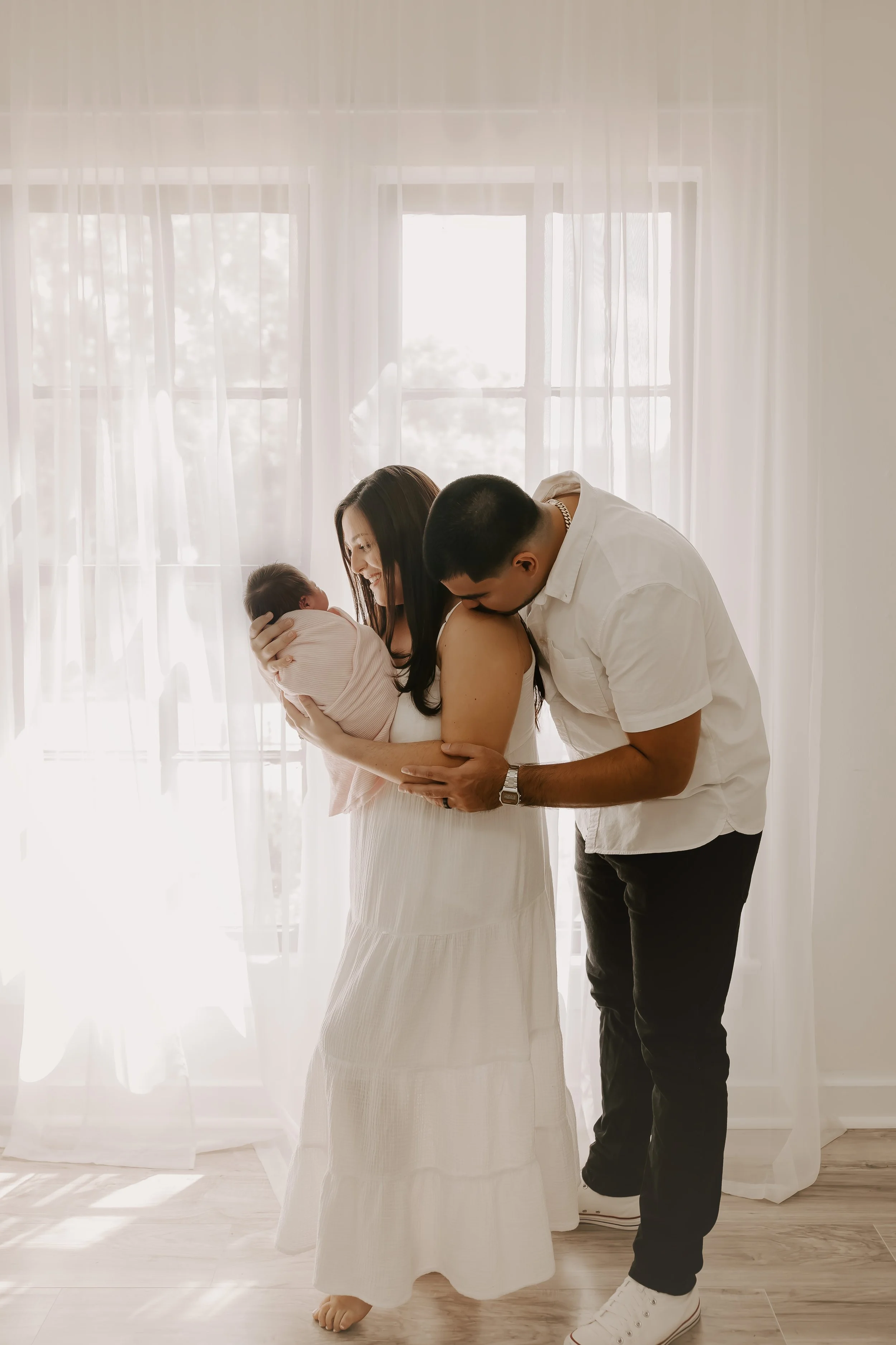 A family in a bright room with white curtains and large windows. The woman holds the baby, and the man is leaning in affectionately.