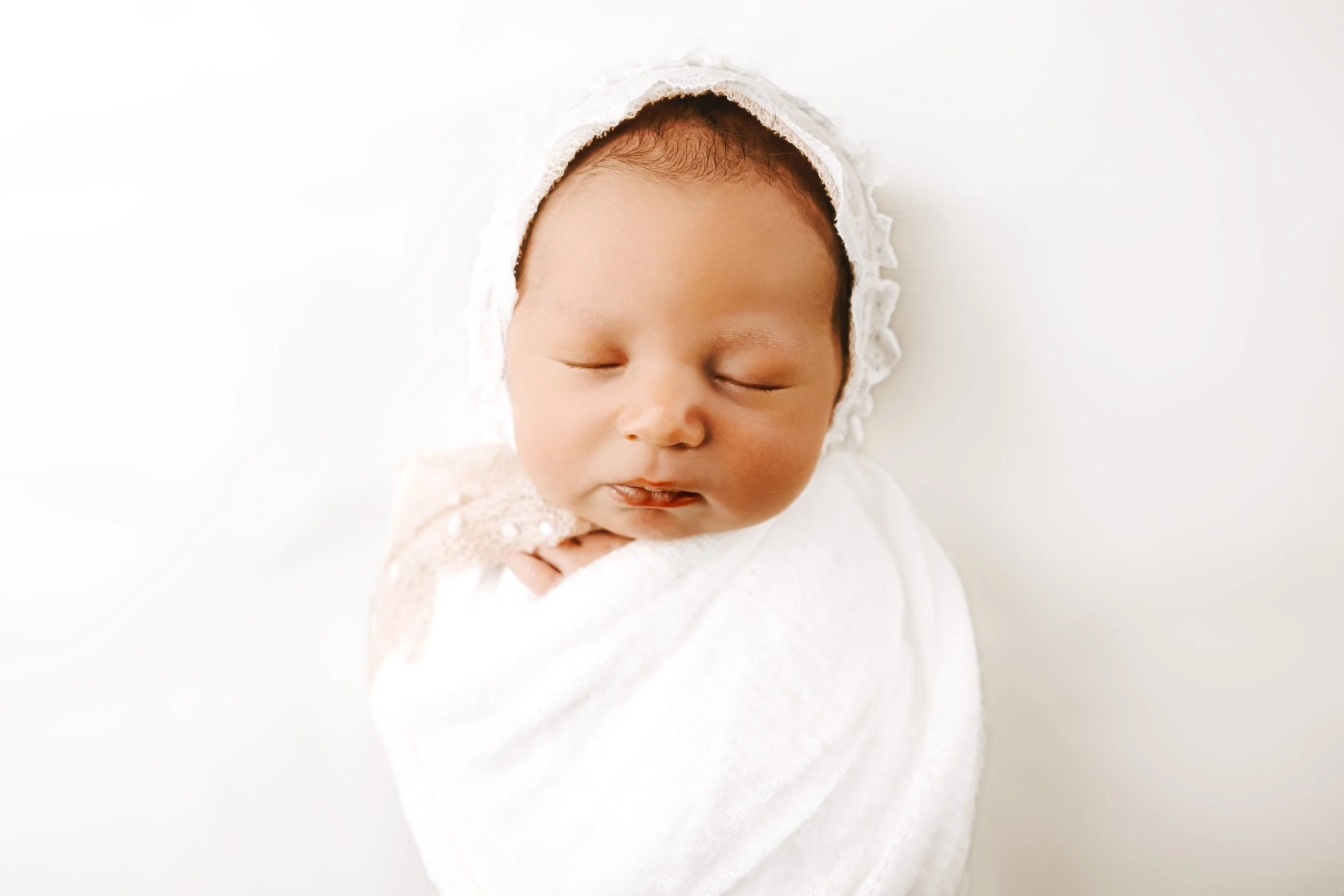Close-up of a sleeping newborn wrapped in a white blanket, wearing a white lace bonnet, with a serene expression against a white background.