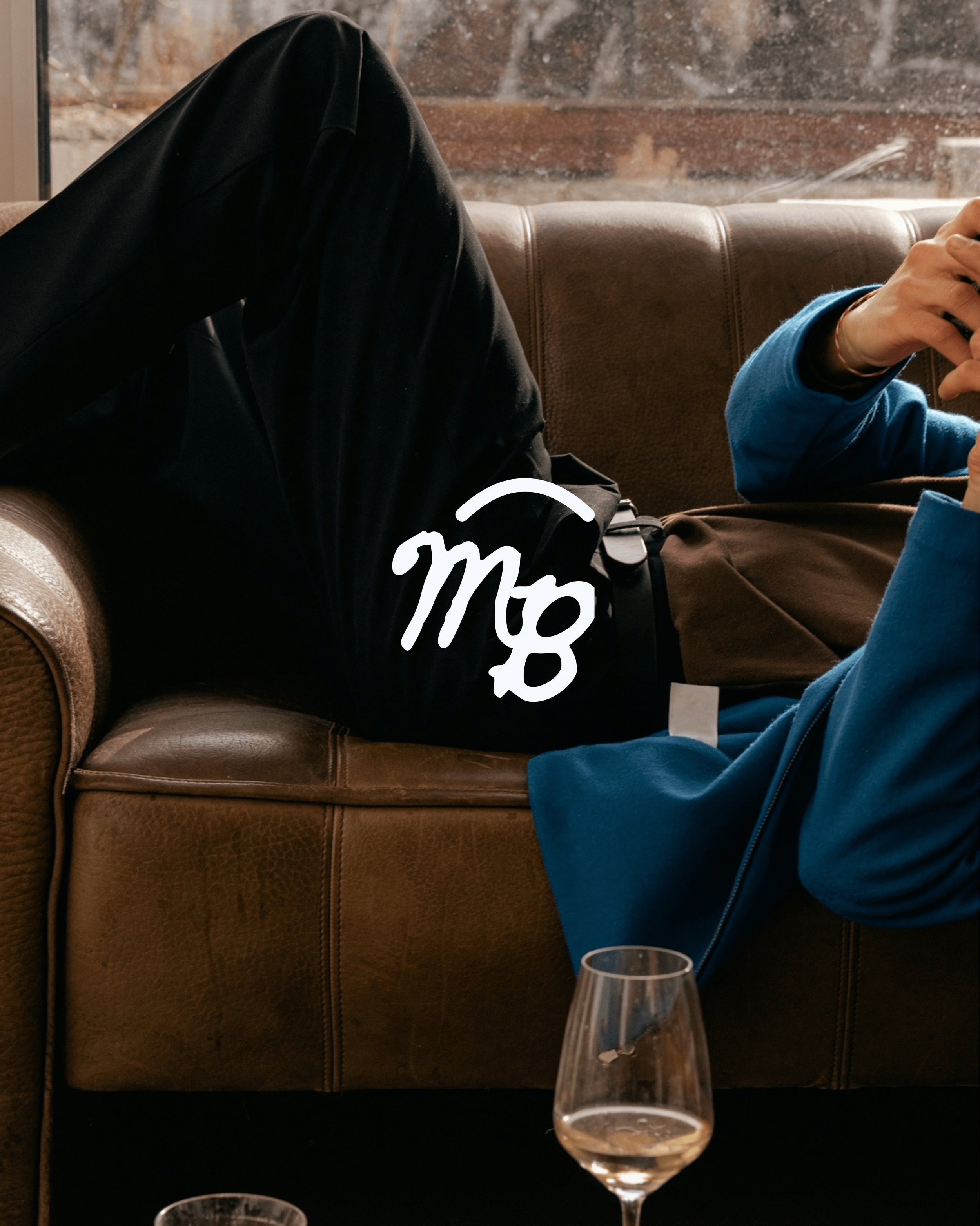 Person lying on a brown leather couch, wearing black pants and a blue jacket, with a white 'mg' logo on the pants. A glass of water is on the table in front of them.