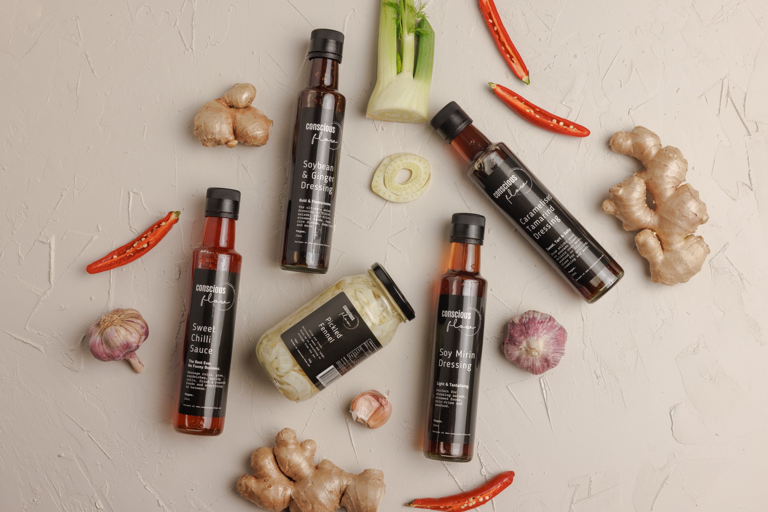 Various bottles of Asian-inspired dressings and condiments, including soybean and ginger, caramelized tamarind, pickled fennel, soy mirin, sweet chili sauce, and garlic, arranged with fresh ingredients.