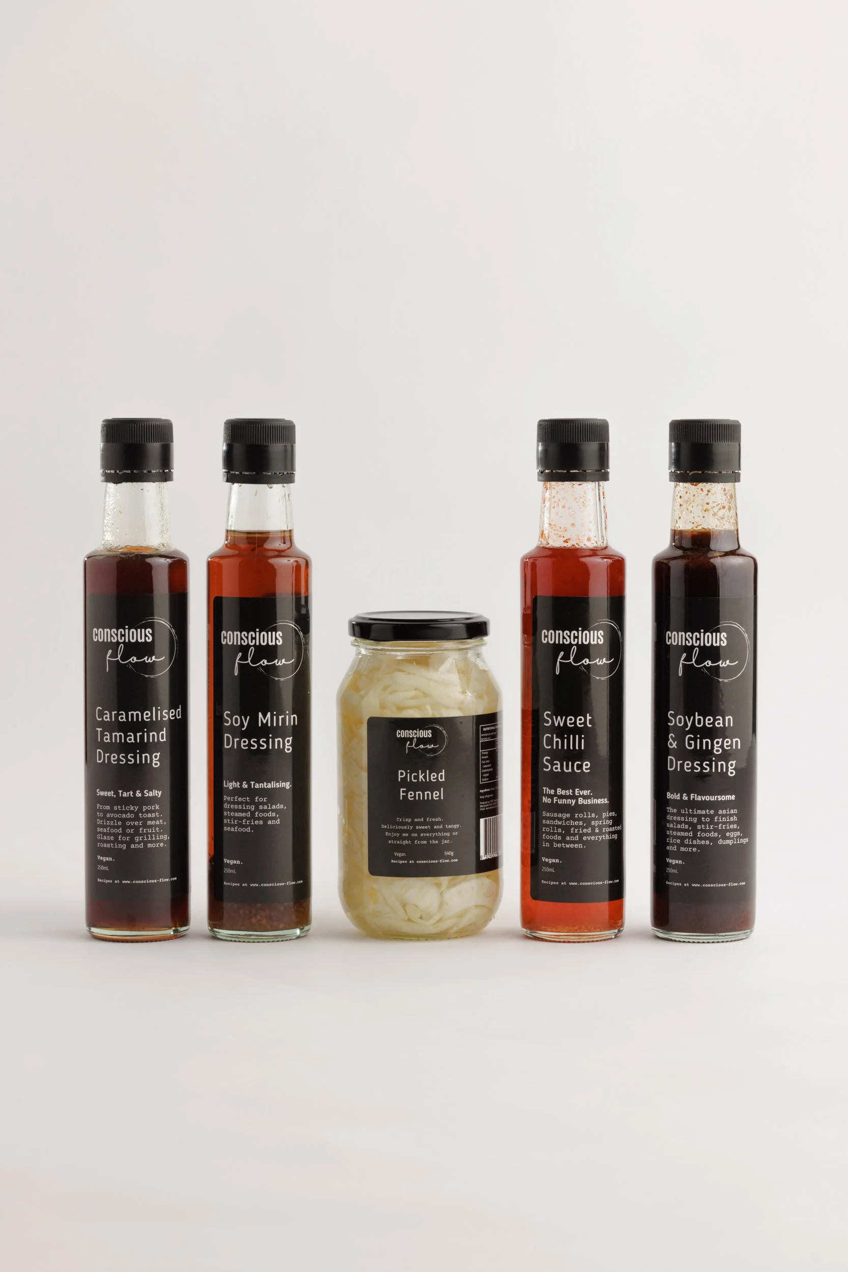 Four bottles of conscious flow dressings, one jar of pickled fennel, all with black caps and labeled in black and white, against a plain white background.