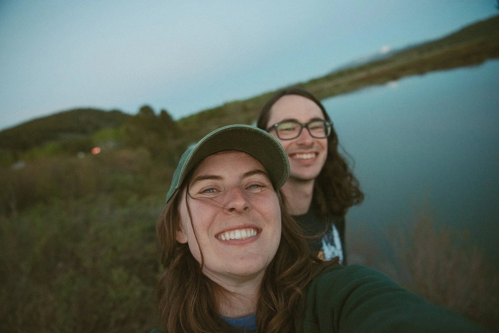 Two friends taking a selfie outdoors near a body of water at dusk, with hills in the background while exploring Grand Teton National Park.