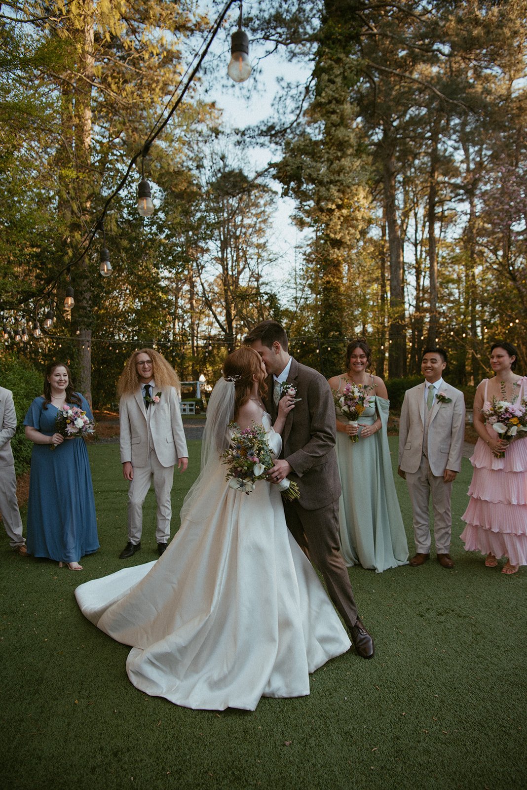 A wedding scene with a bride and groom kissing, surrounded by bridesmaids and groomsmen outdoors on a grassy area with trees and string lights.
