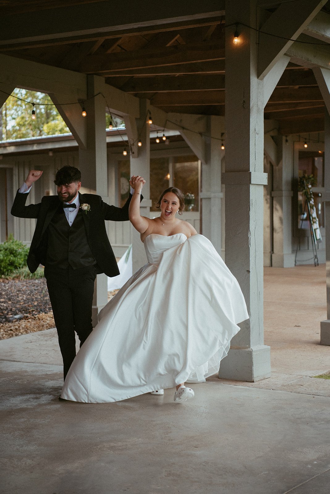 A bride and groom celebrating, holding hands and smiling, outdoors near a rustic wooden building with string lights overhead.