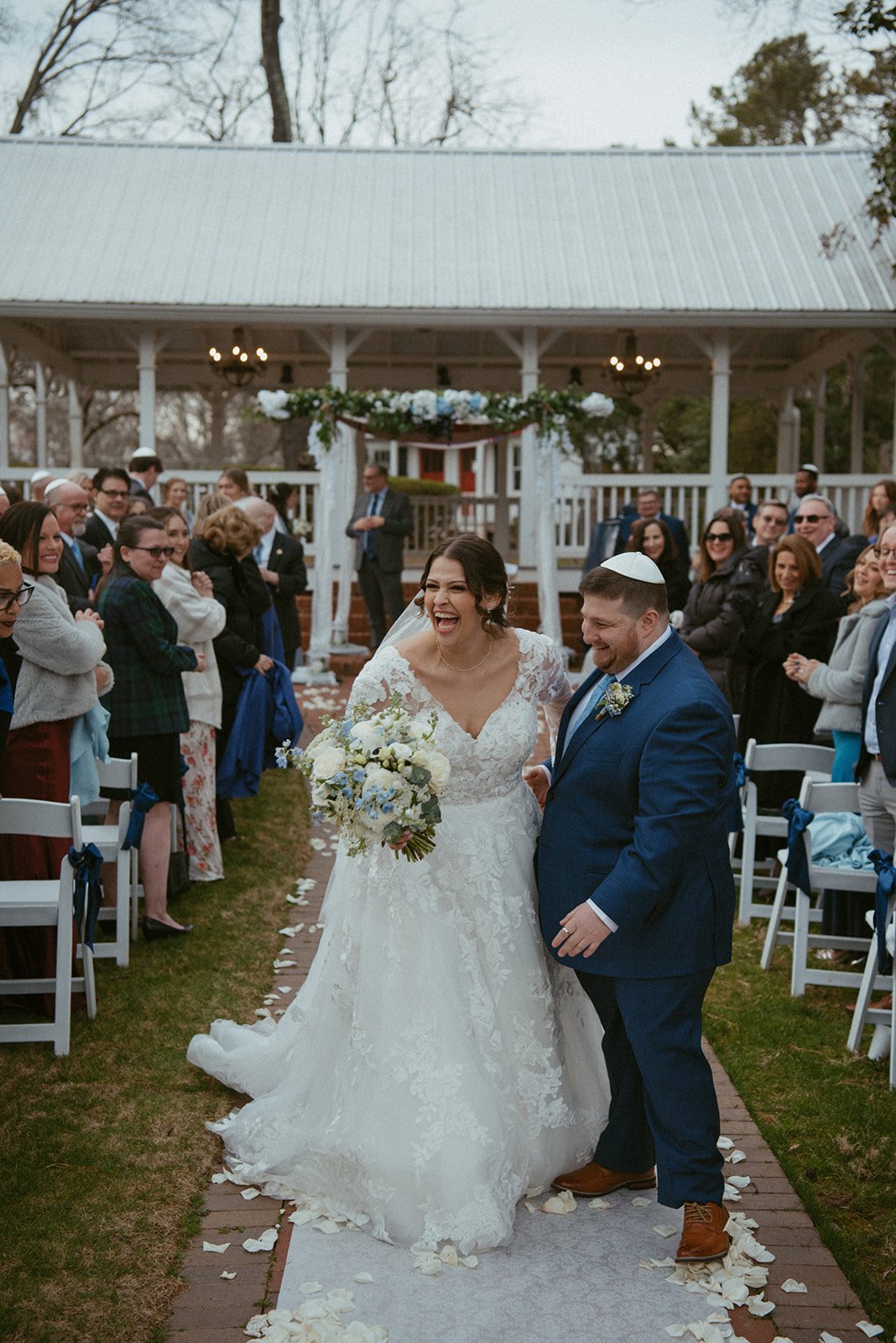 A bride and groom smiling and walking down the aisle at their wedding ceremony, surrounded by friends and family outside.
