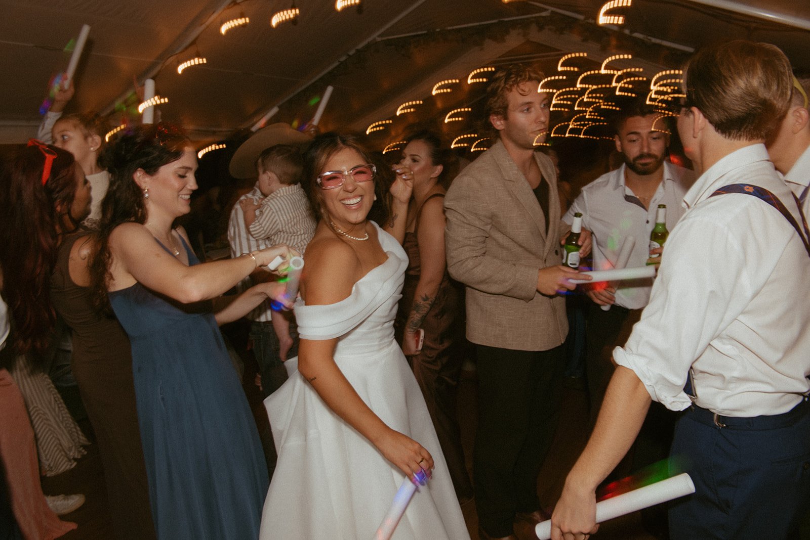 Group of people celebrating at a lively party, some holding glow sticks, with smiling and laughing expressions, under warm lighting.