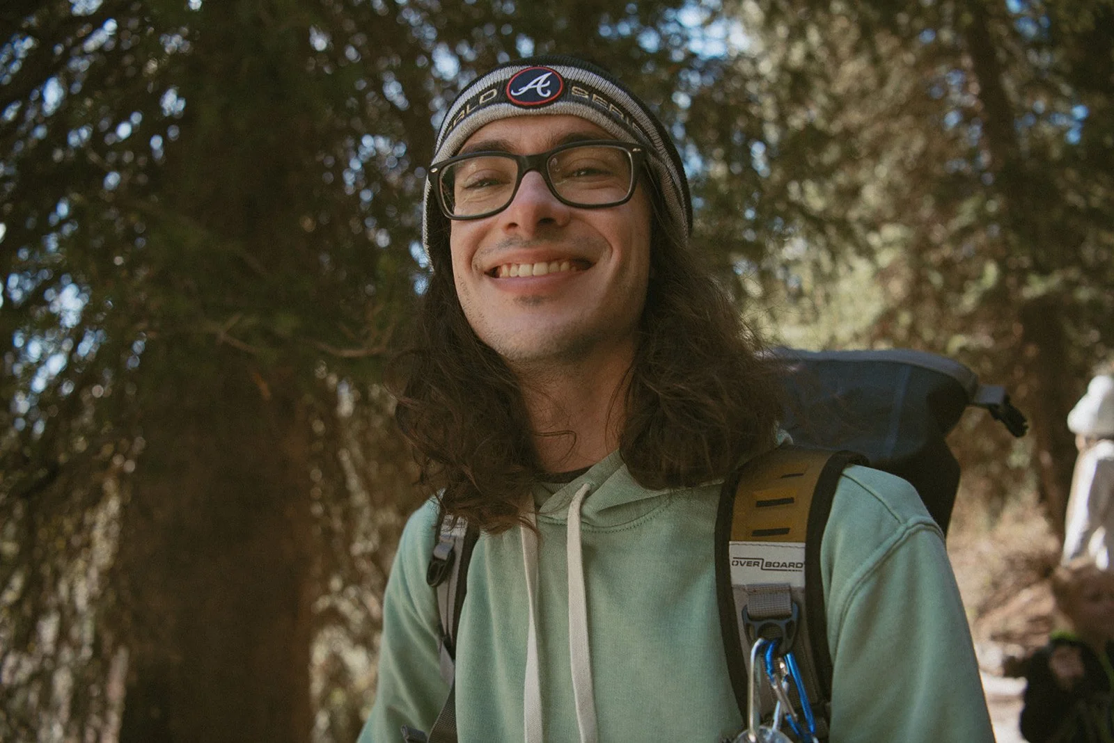 A smiling man with long hair, glasses, wearing a beanie, hoodie, and backpack outdoors in a wooded area.