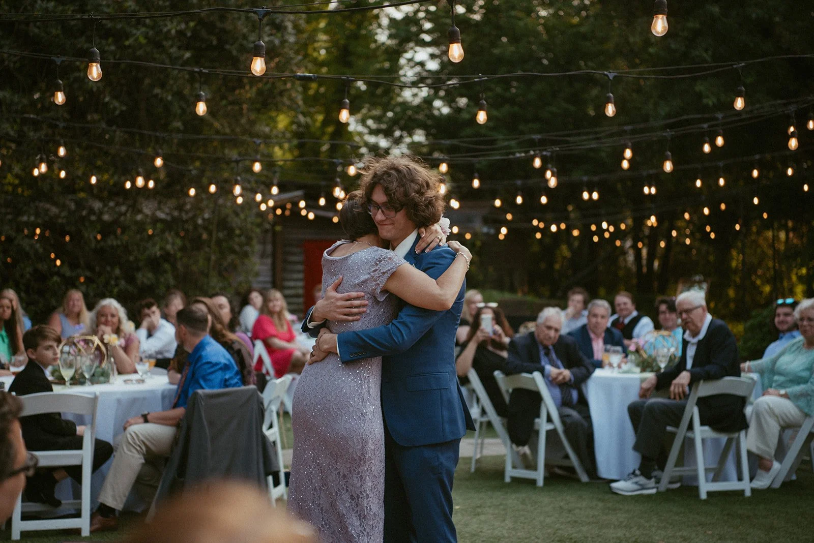 A mother and a young man dance together at an outdoor evening wedding reception. The scene is decorated with string lights, and guests are seated at tables in the background.