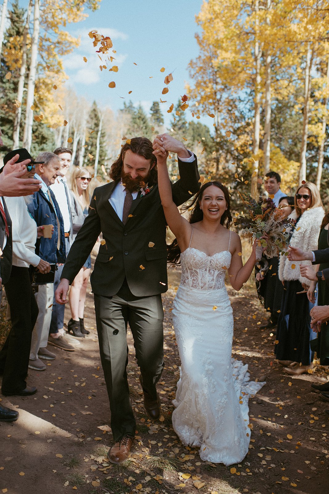 Happy couple, likely newlyweds, walking through a tree-lined outdoor area during the fall, throwing leaves and smiling, with guests on either side celebrating.