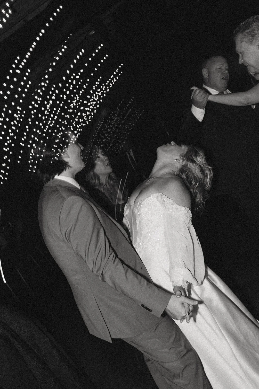 A black and white photo of a wedding dance with the bride and groom holding hands and looking into each other's eyes, surrounded by guests, under a ceiling decorated with string lights.