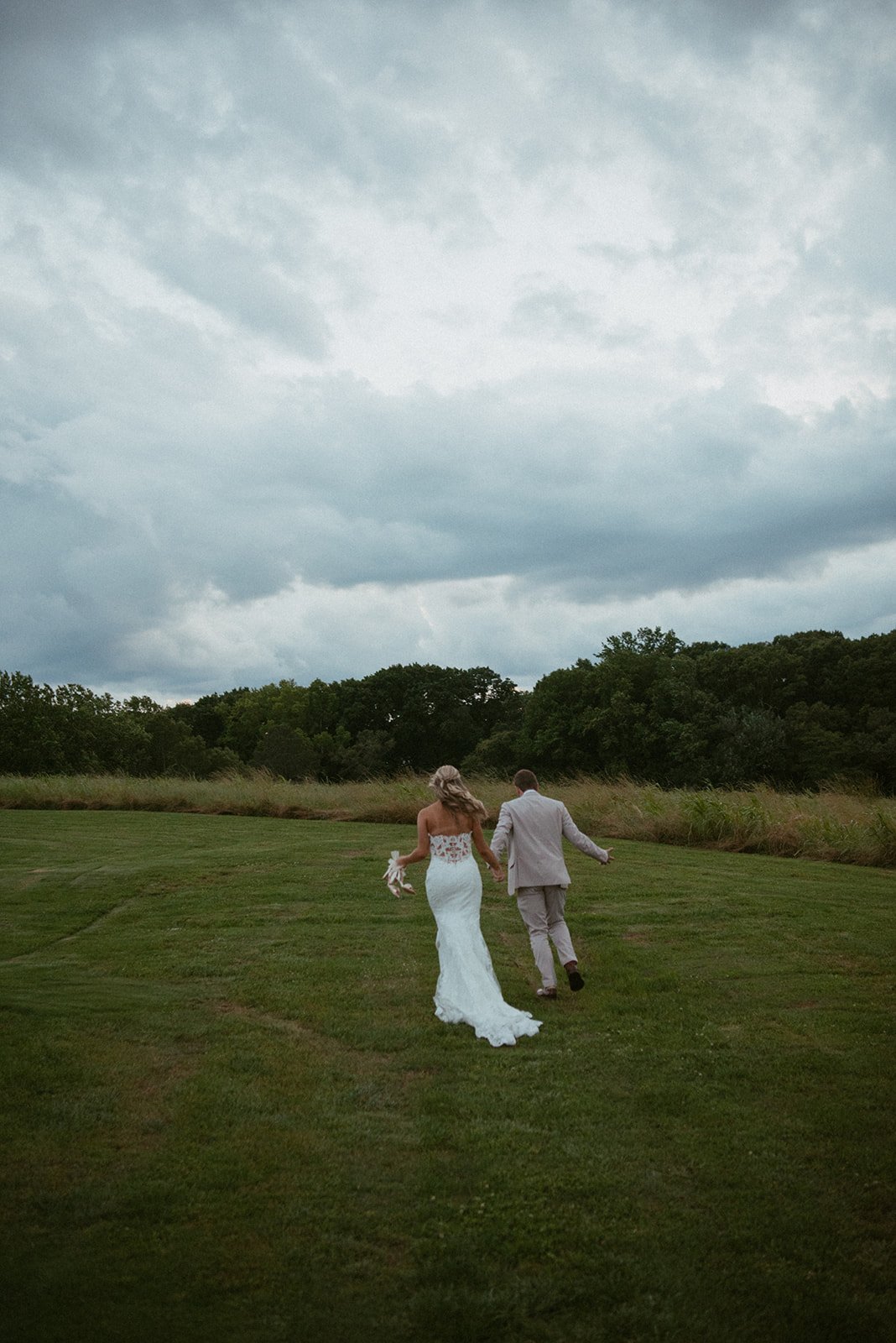 Bride and groom walking hand in hand across a grassy field before a cloudy sky, with trees in the background.