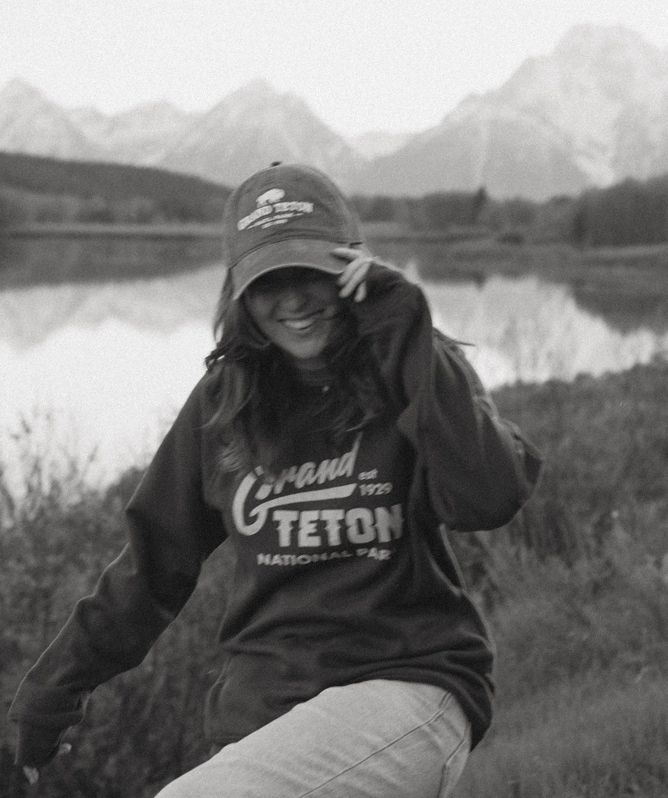 A woman smiling outdoors at a lake with mountains in the background, wearing a baseball cap and a hoodie that says 'Glam Teton'.