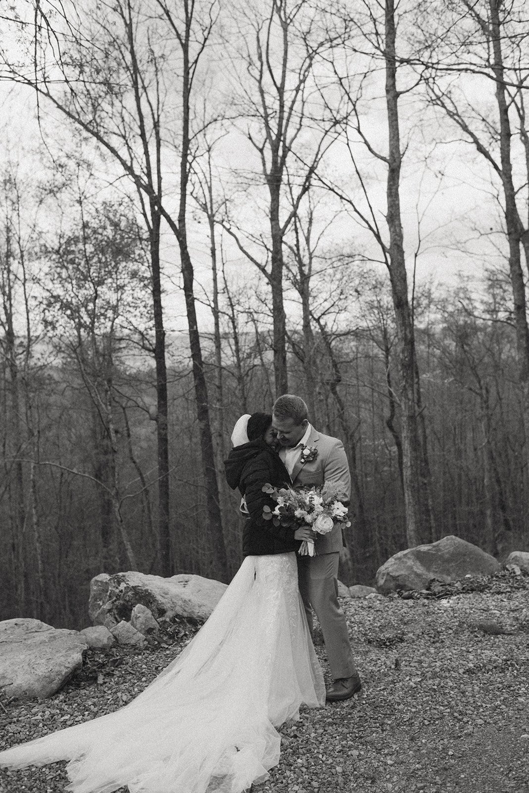 A black and white photo of a bride and groom hugging outdoors in a wooded area. The bride wears a long wedding dress and holds a bouquet, and the groom wears a suit with a boutonniere. They are embracing each other.