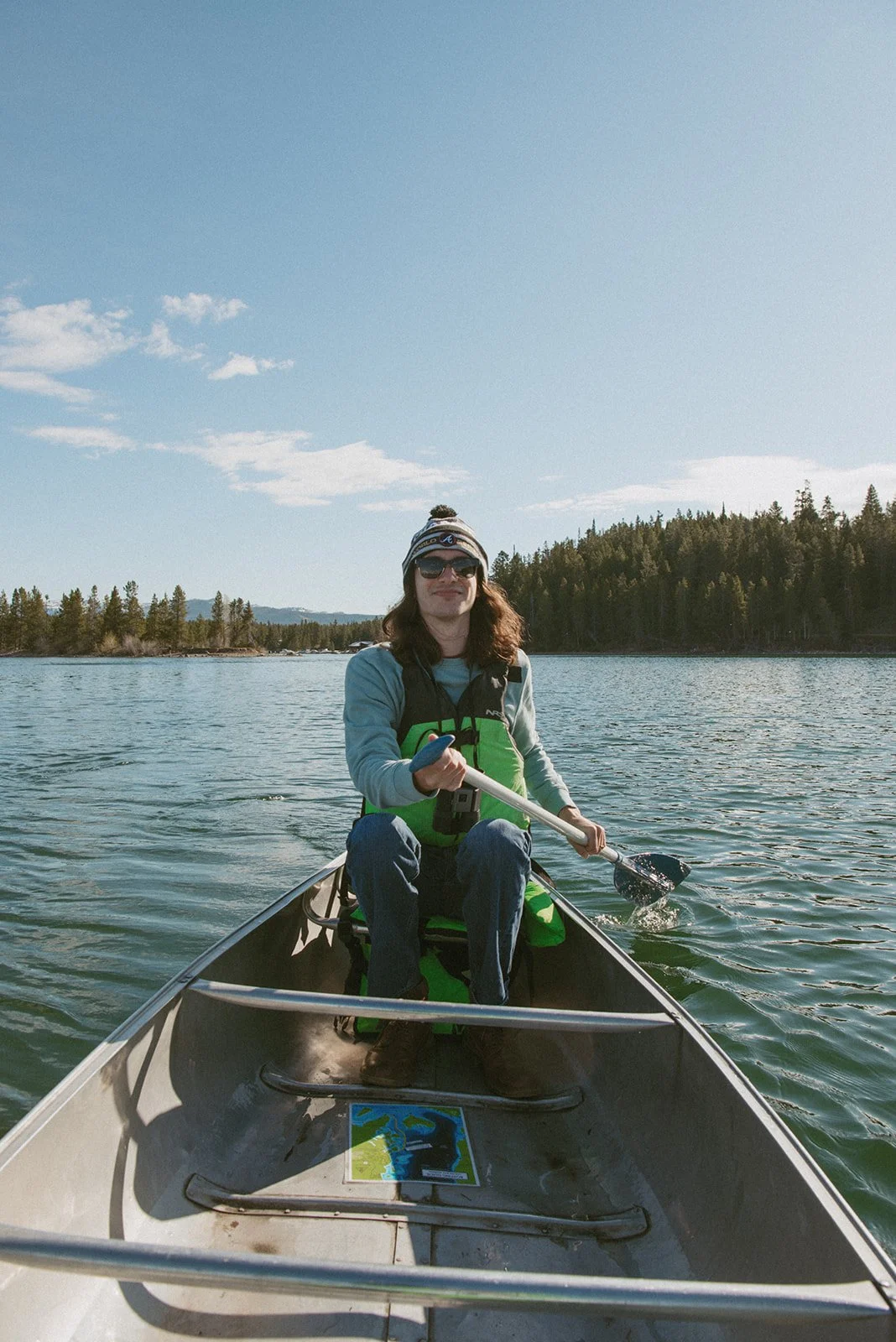 Person kayaking on a lake surrounded by trees, wearing sunglasses, a hat, and a life jacket, with a map visible inside the kayak.