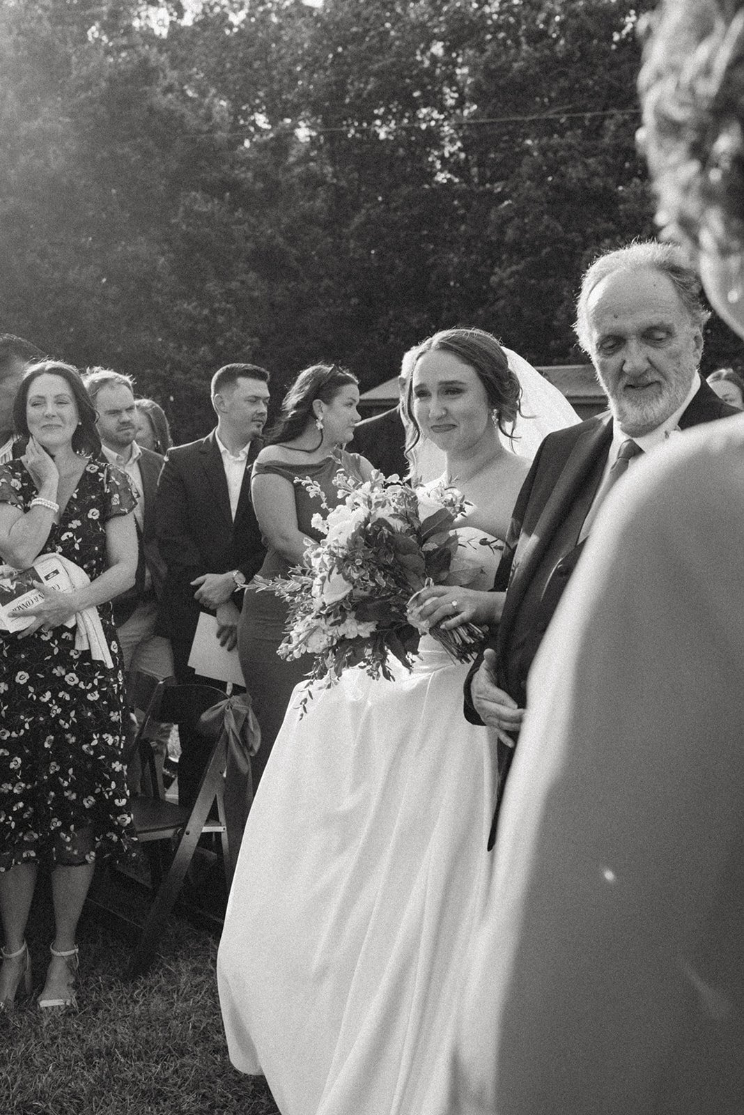 A black and white photo of a bride holding a bouquet of flowers, standing next to an older man in a tuxedo at an outdoor wedding ceremony, with several guests in the background.