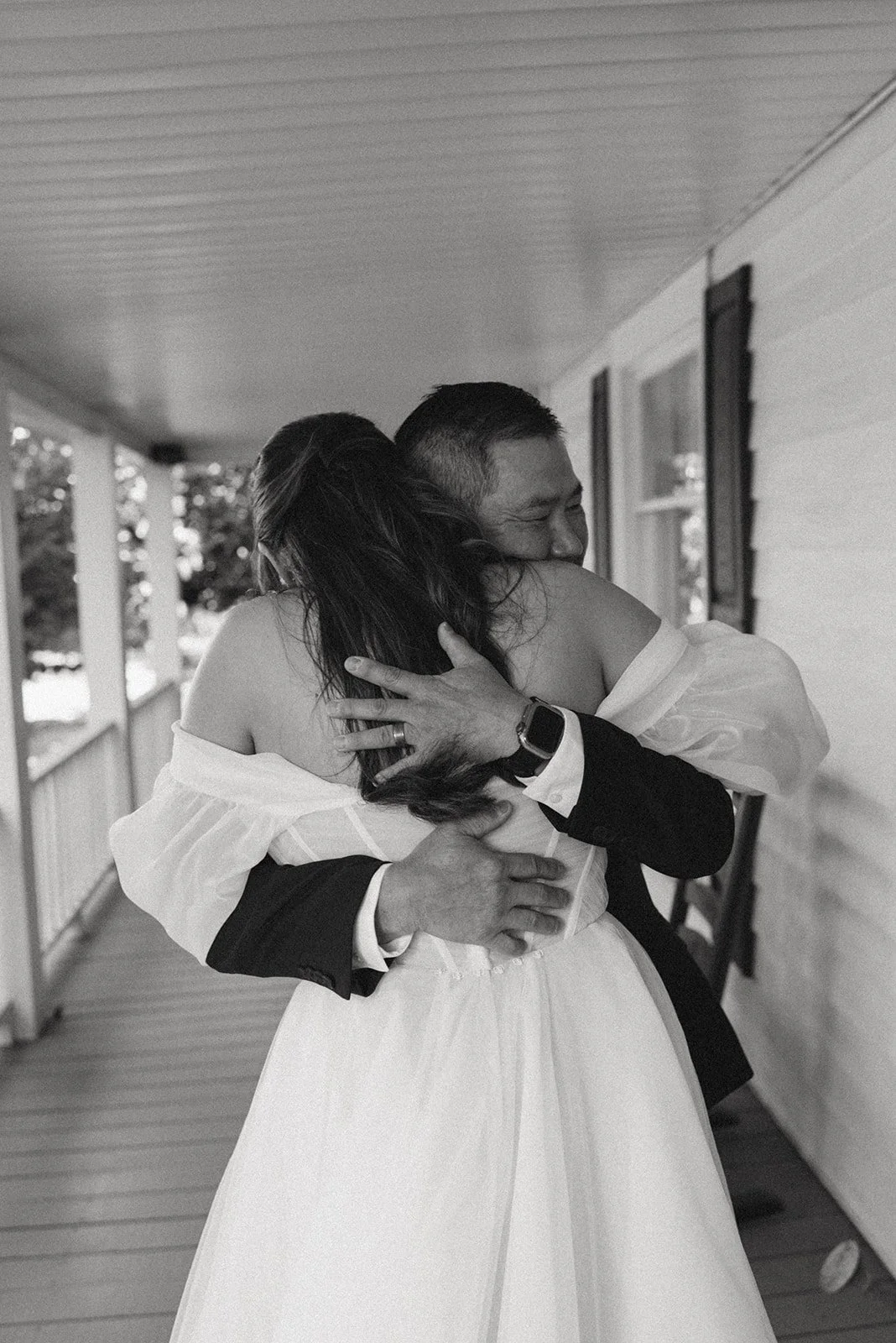 Black and white photo of a couple embracing on a porch, the woman in a wedding dress and the man in a suit, showing happiness and affection.