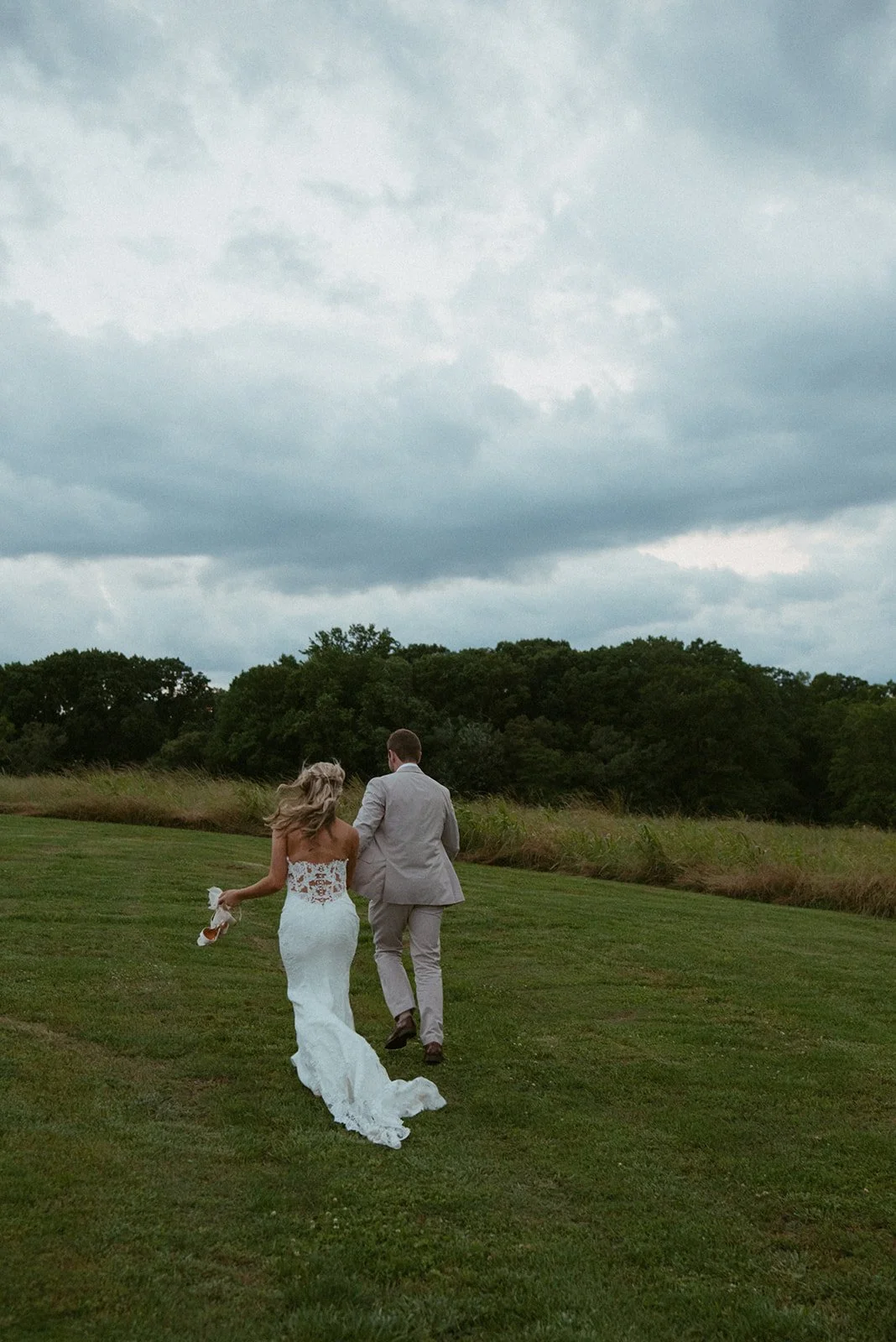 A newlywed couple walking across a grassy field with trees in the background under a cloudy sky, the woman in a white lace wedding dress and the man in a light grey suit.