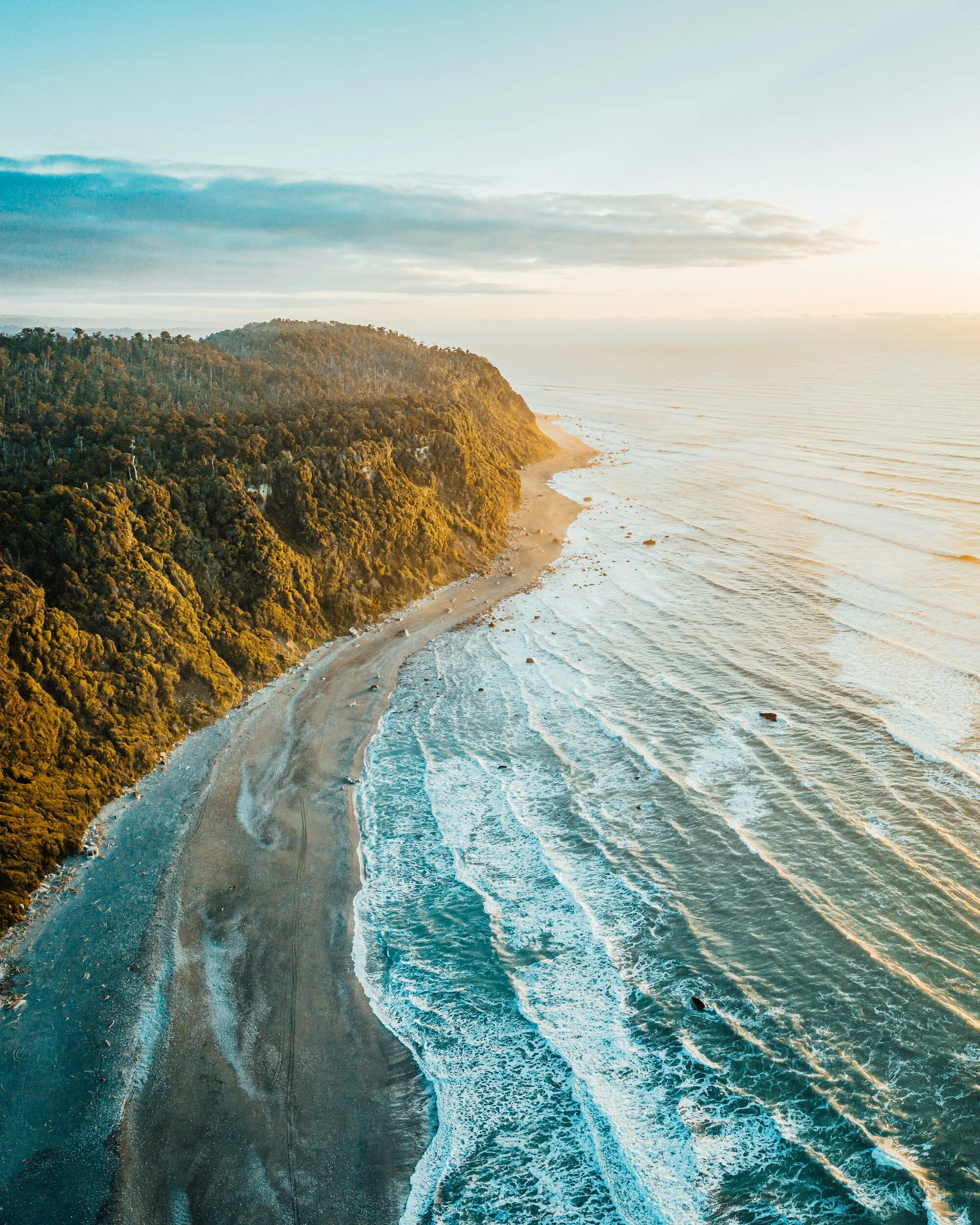 Aerial view of a coastline with green forested cliffs, sandy beach, and ocean waves during sunset.