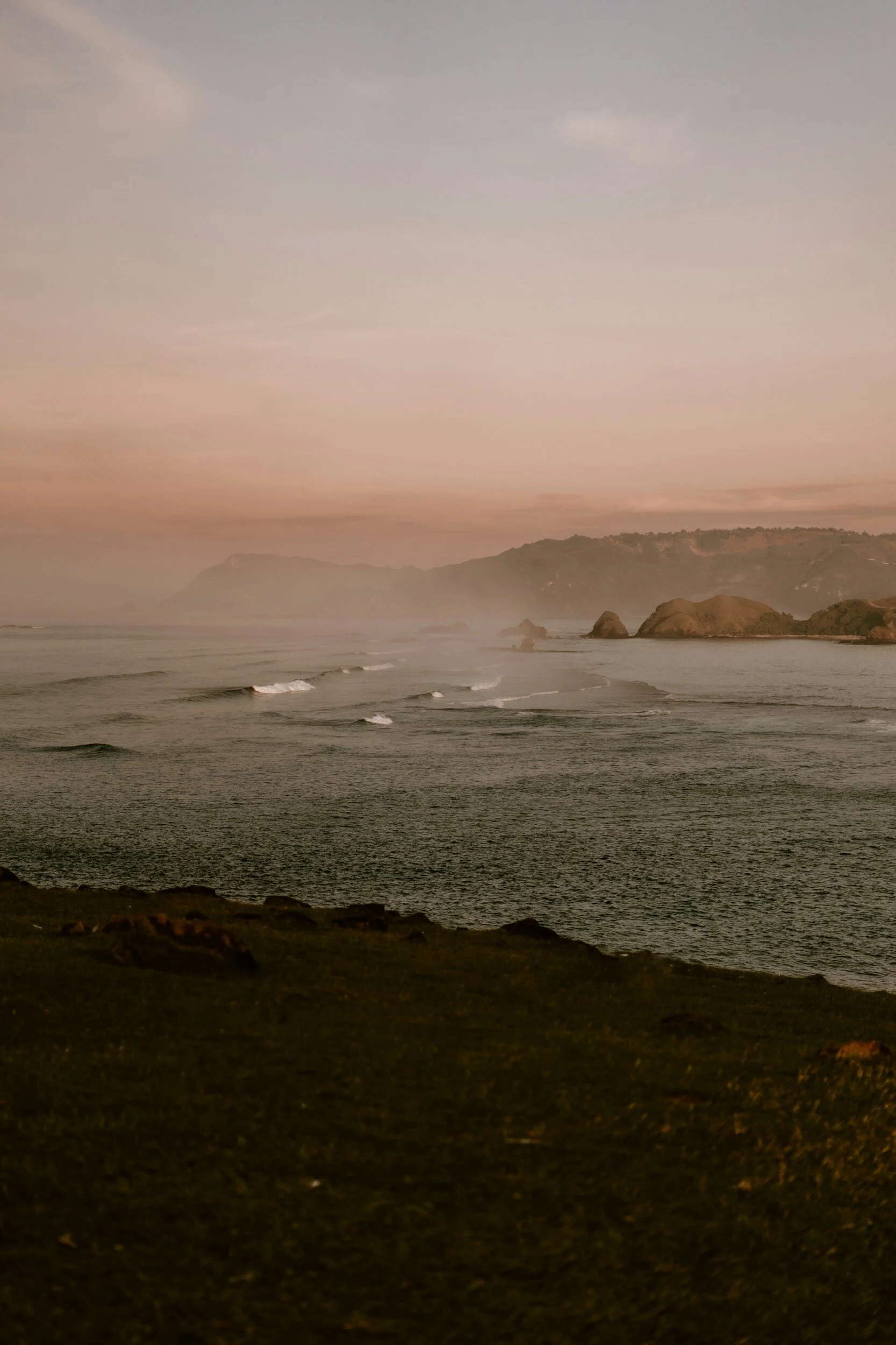 A coastal landscape at sunset with waves hitting the shoreline, distant hills, and rocky formations extending into the ocean under a pastel-colored sky.