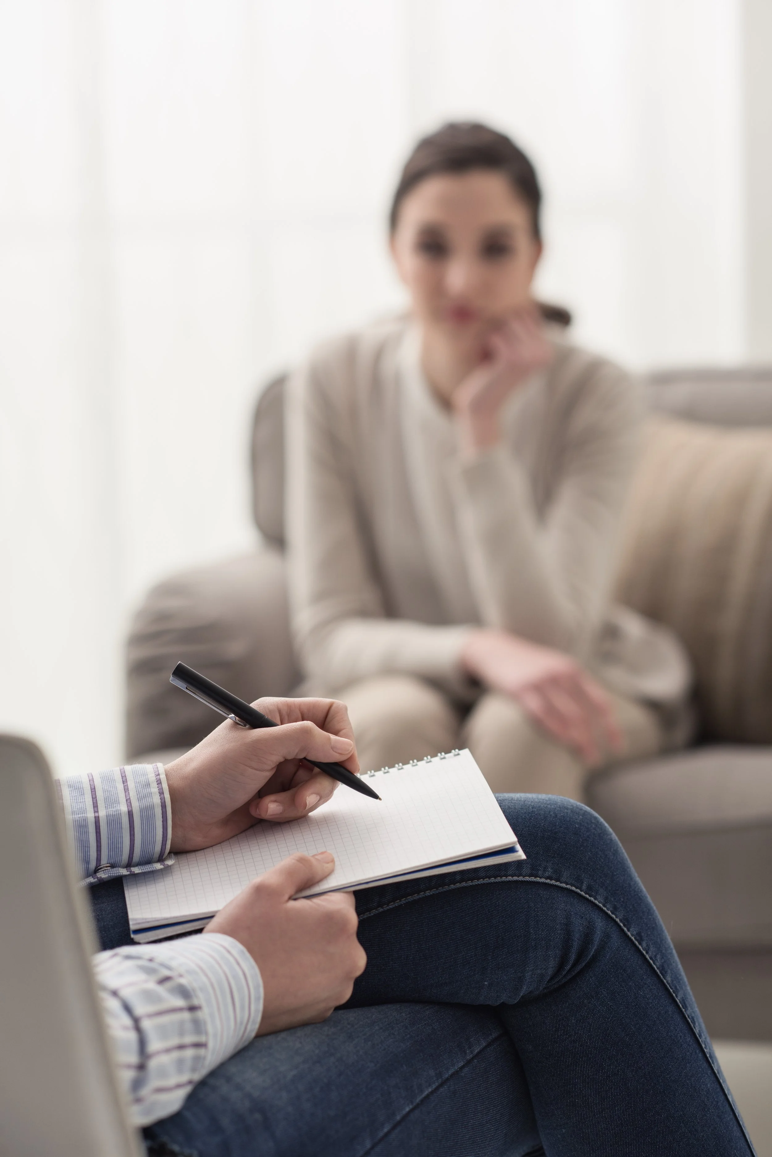 A woman sitting on a couch, blurred in the background, while a person in the foreground takes notes on a notepad.