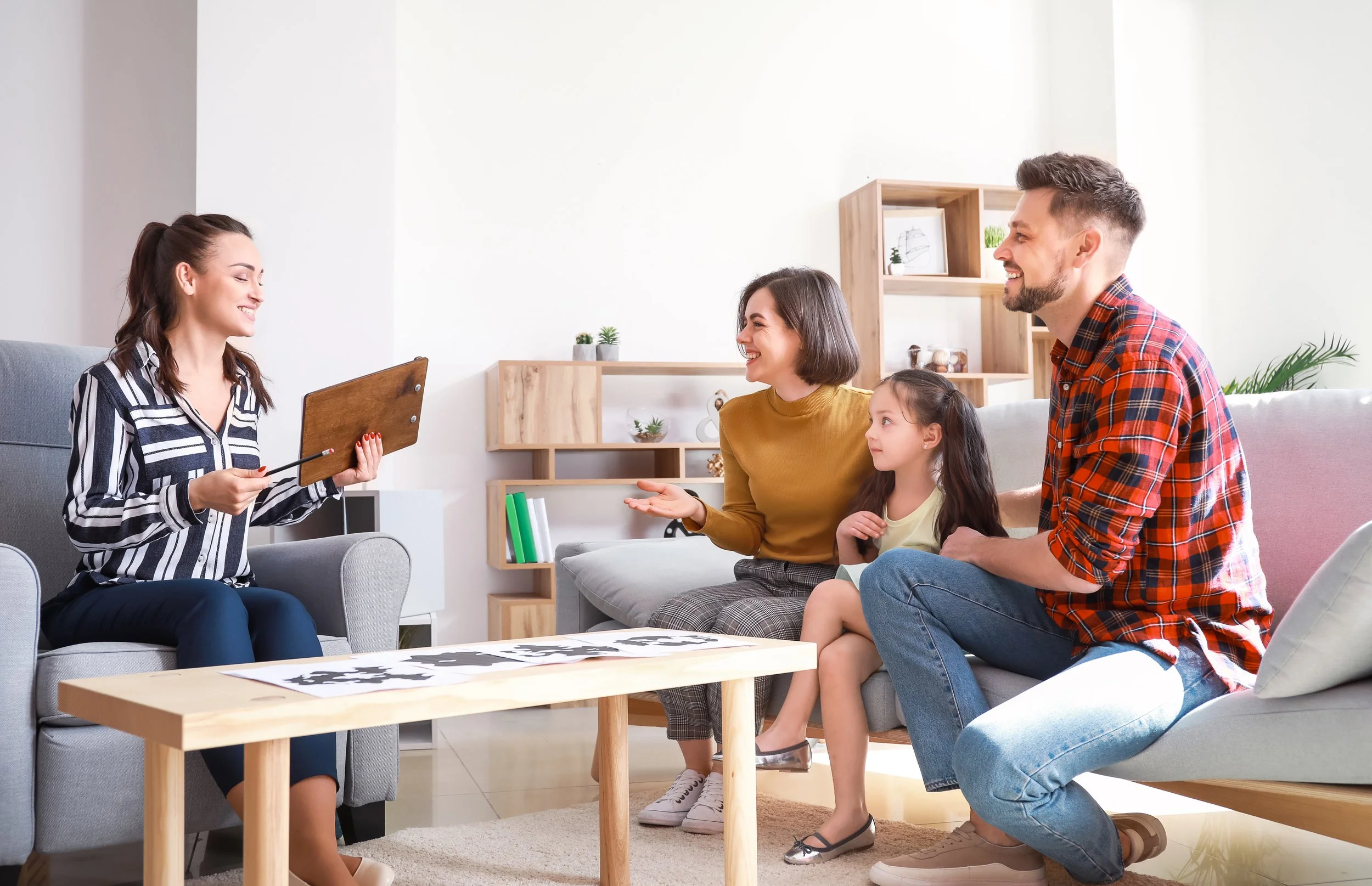 A woman sitting on a sofa showing art on a clipboard to a family of four. Family members are smiling; the woman on the sofa wearing a mustard sweater, the man in a red plaid shirt, a young girl in a light yellow shirt, and a woman in a striped blouse holding a piece of art on paper. The room has shelves with books and decorations, with natural light coming in.