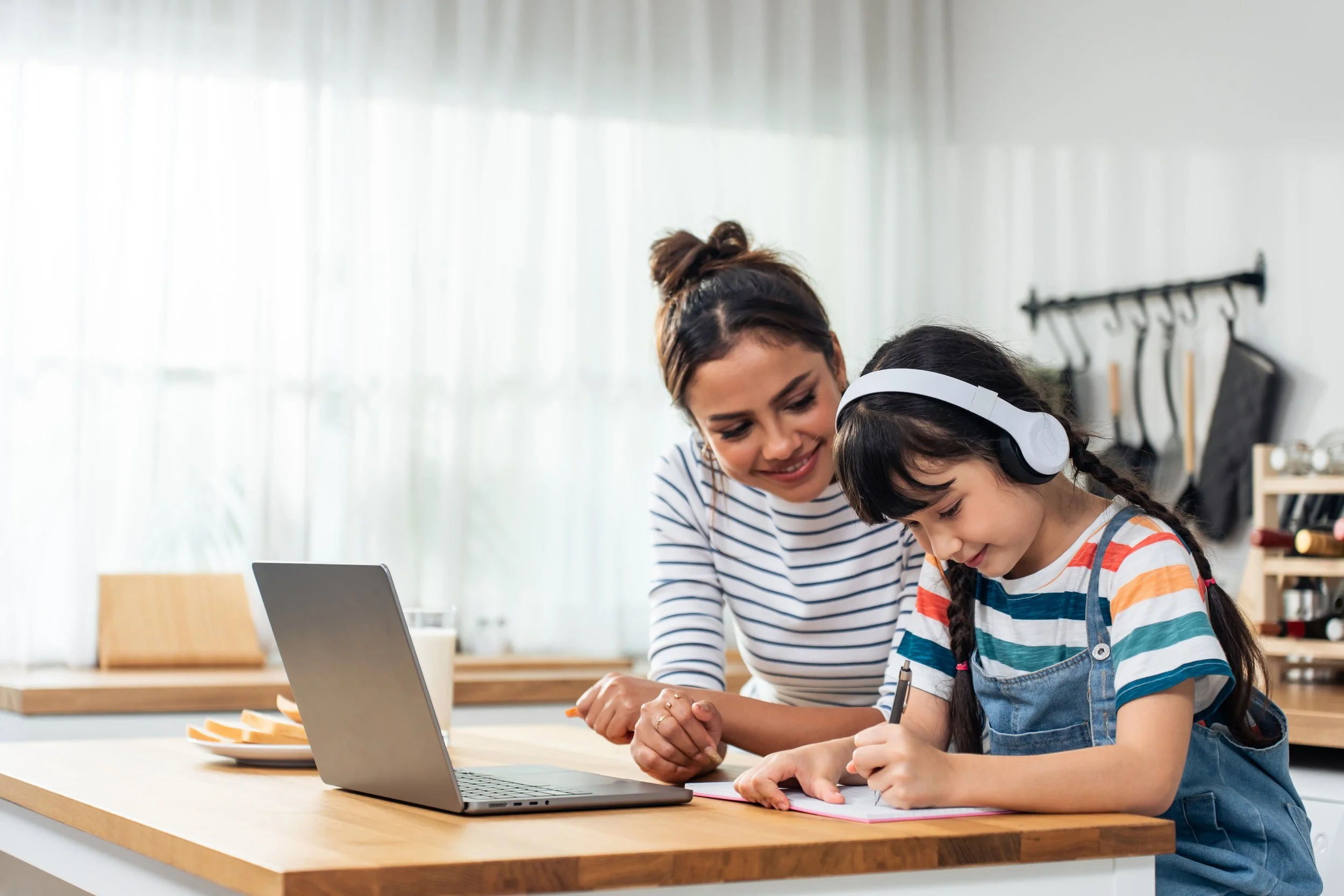 A woman and a young girl with headphones at a kitchen island, working together with a laptop and notebook.