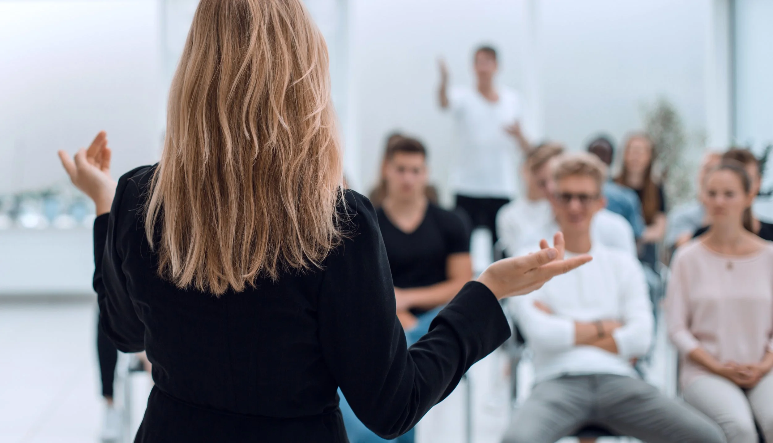 Woman with blonde hair giving a presentation to a group of diverse young adults sitting in a bright room with large windows.