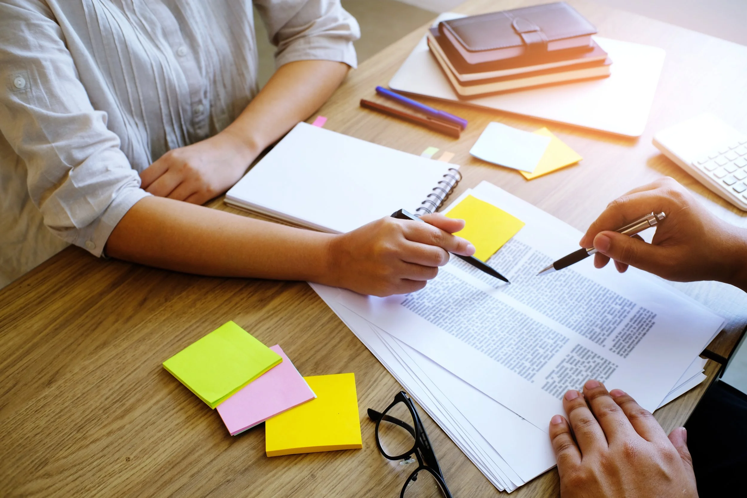 Two people discussing documents and taking notes on a wooden table with sticky notes, notebooks, pens, glasses, and a keyboard.