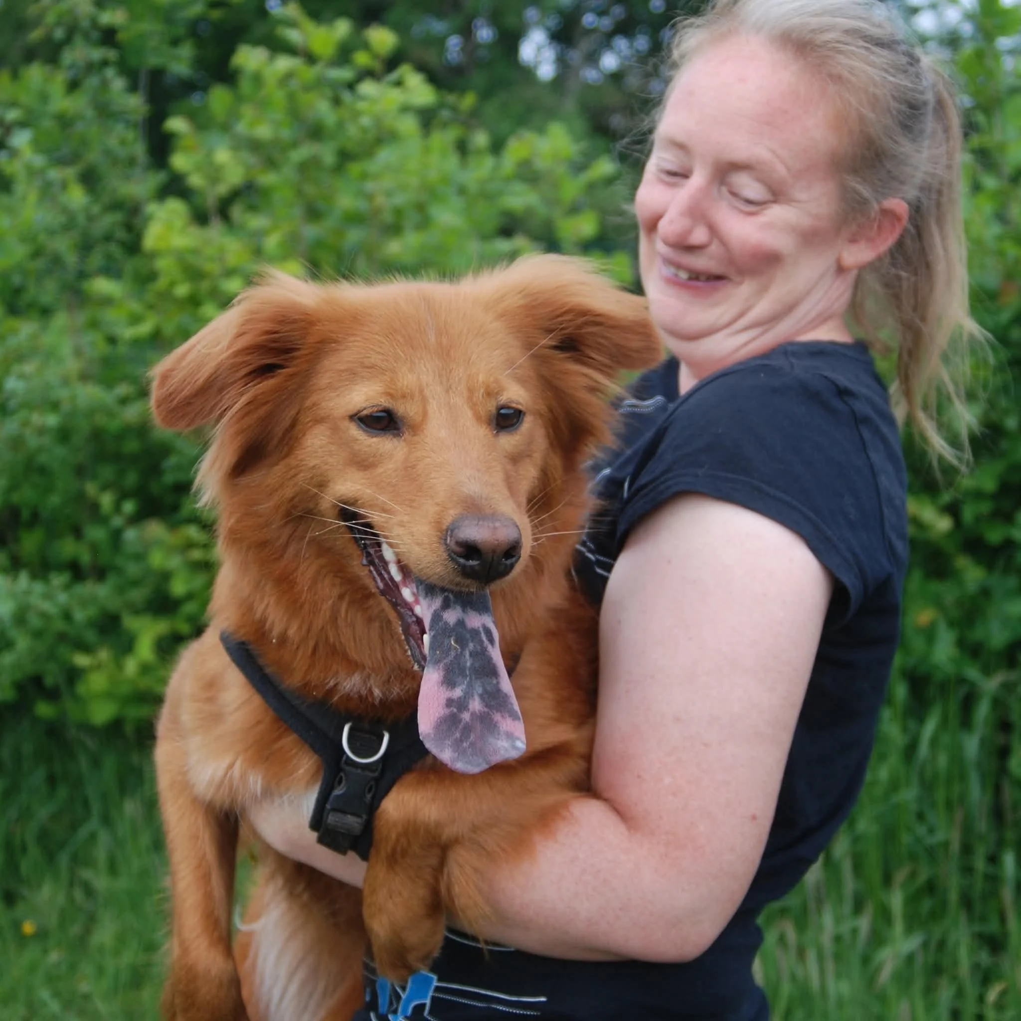 A white woman with light brown hair, smiling with a ginger dog with black spots on their tongue