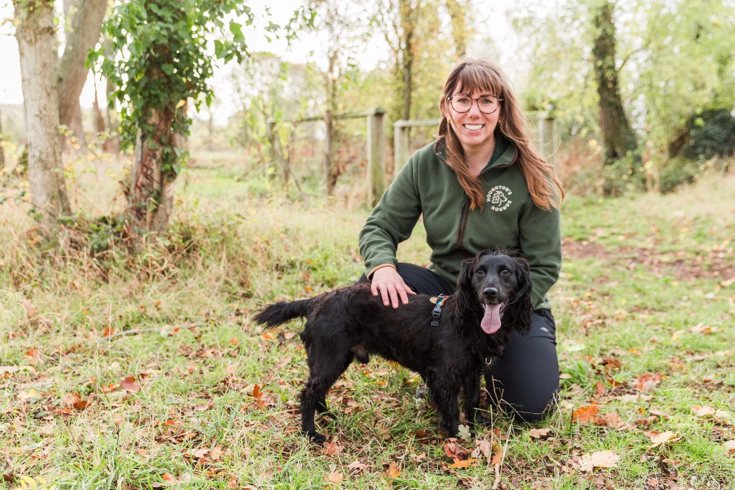 A woman wearing glasses and smiling crouched down with a black cocker spaniel in front of her 