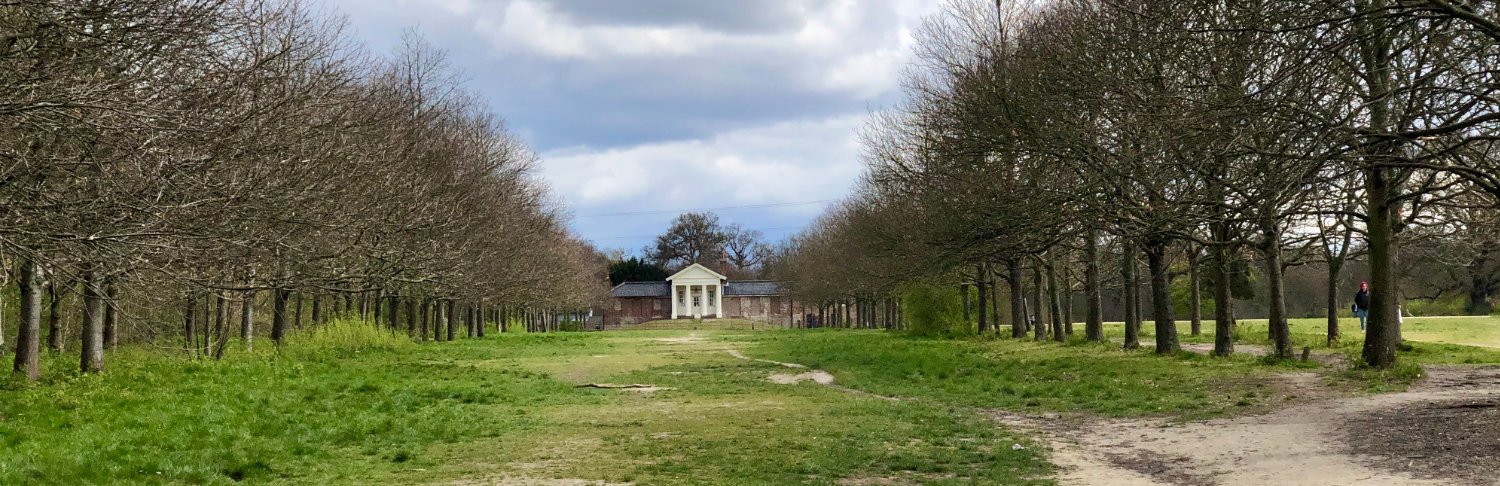 A historic brick stable block building next to leafy green trees and open parkland near Wanstead Park.