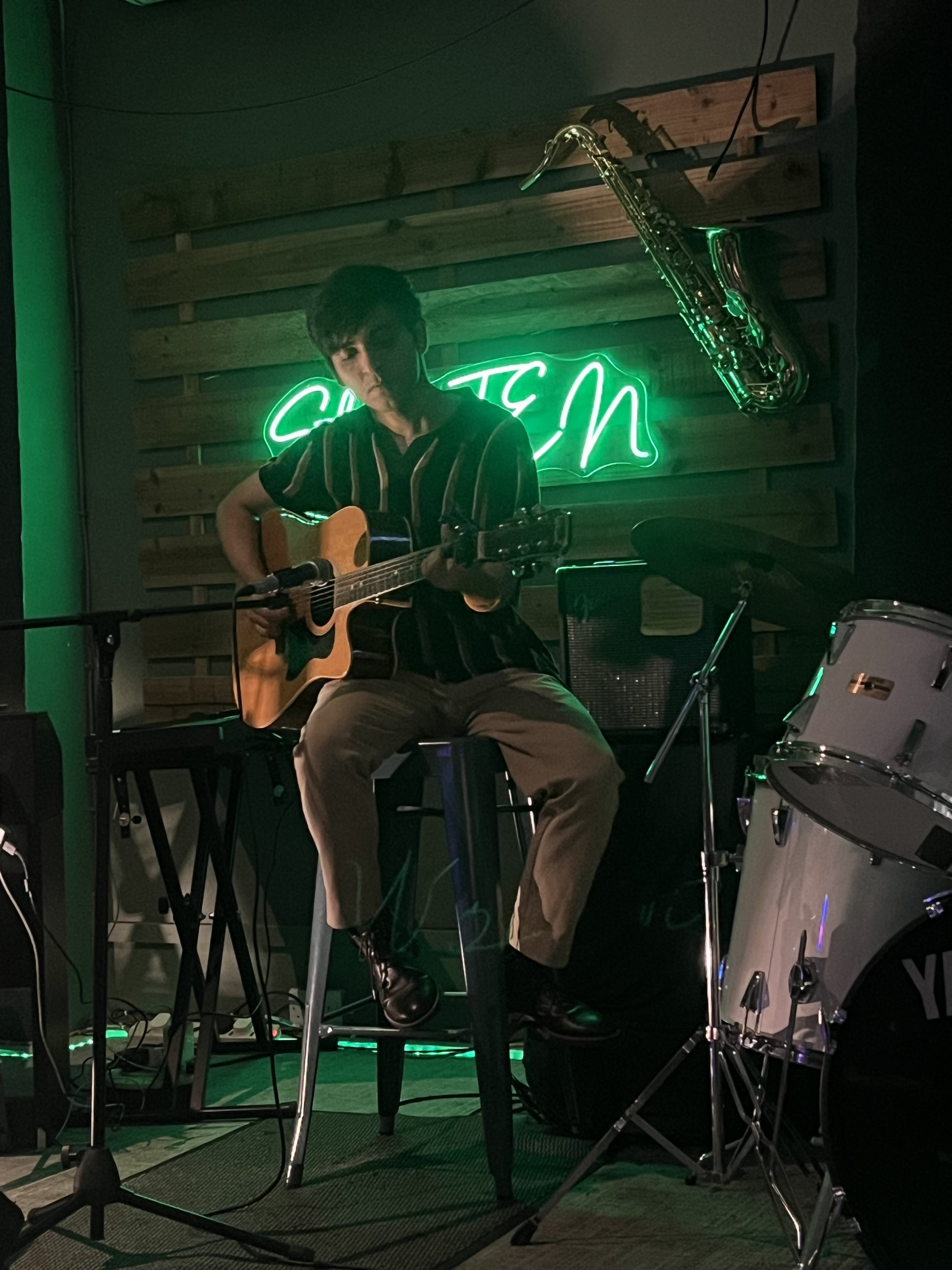 A young man playing an acoustic guitar and singing on stage, with a neon green 'STATION' sign and a saxophone hanging on the wooden panel behind him, accompanied by a drum set and a microphone stand.