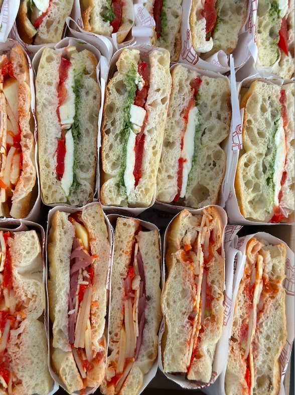 Multiple sandwiches in paper wrappers with fillings like basil cream, tomato, cheese, and meats, stacked closely together.