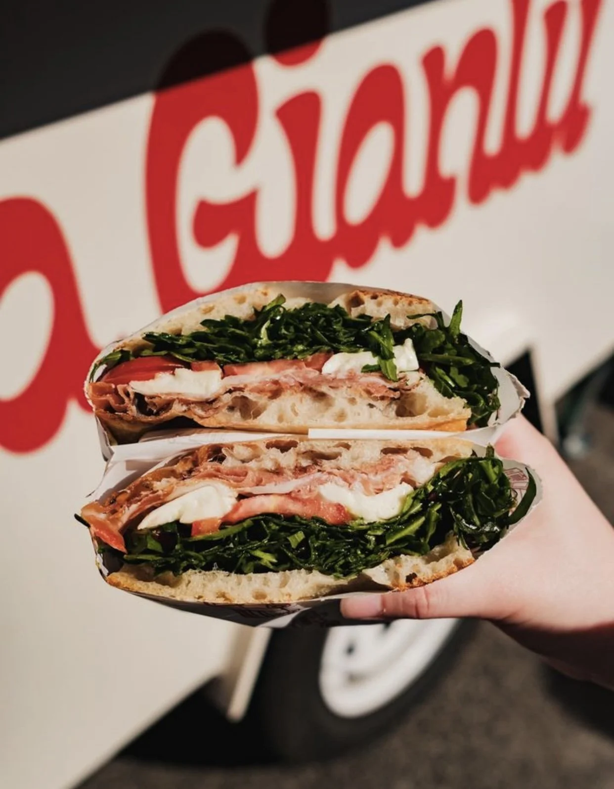 A hand holding a sandwich with greens, tomato, cheese, and meat, in front of a red and white sign.