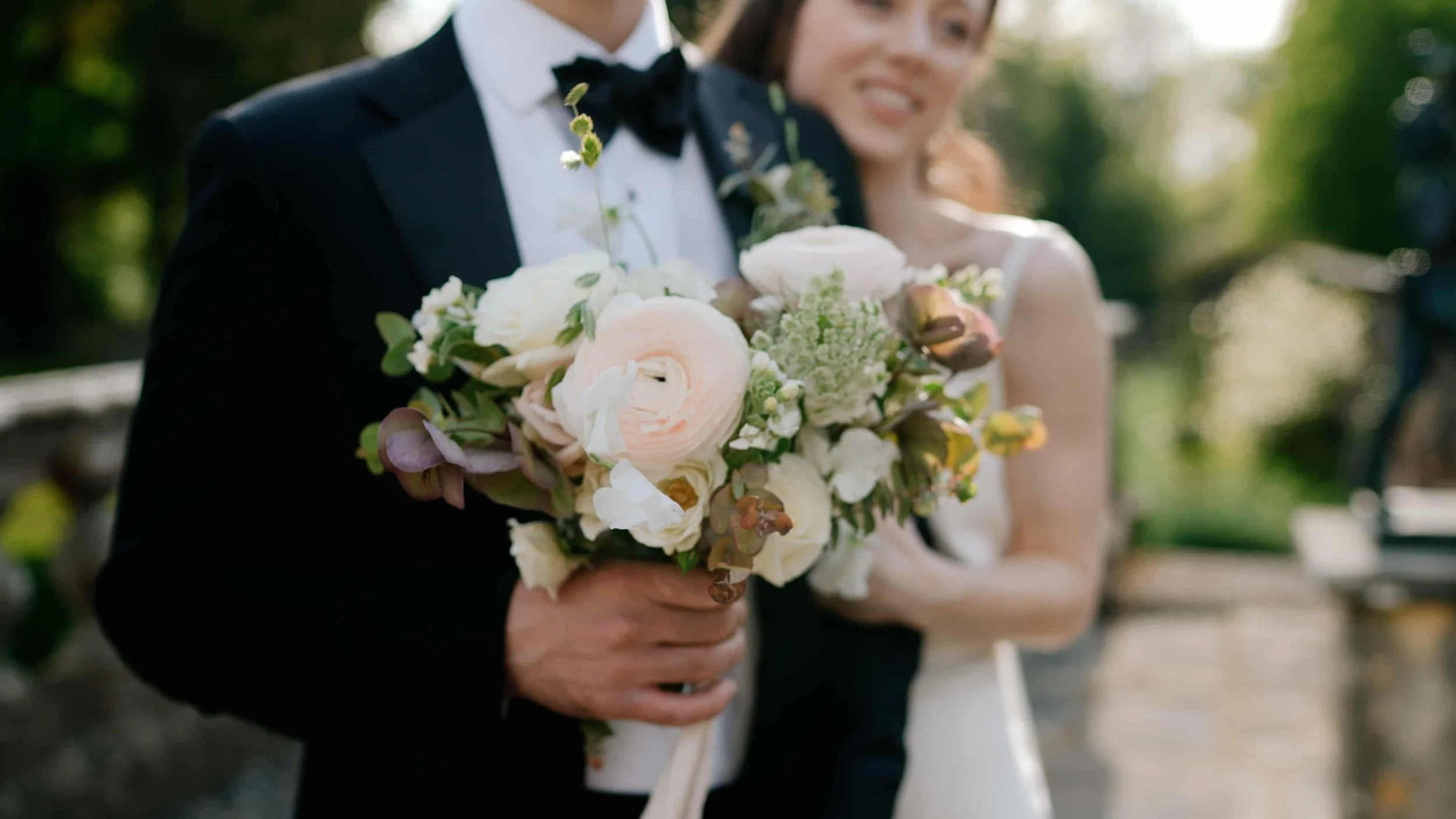 Groom holds brides bouquet while his bride hugs him from behind in the gardens at Gloster House