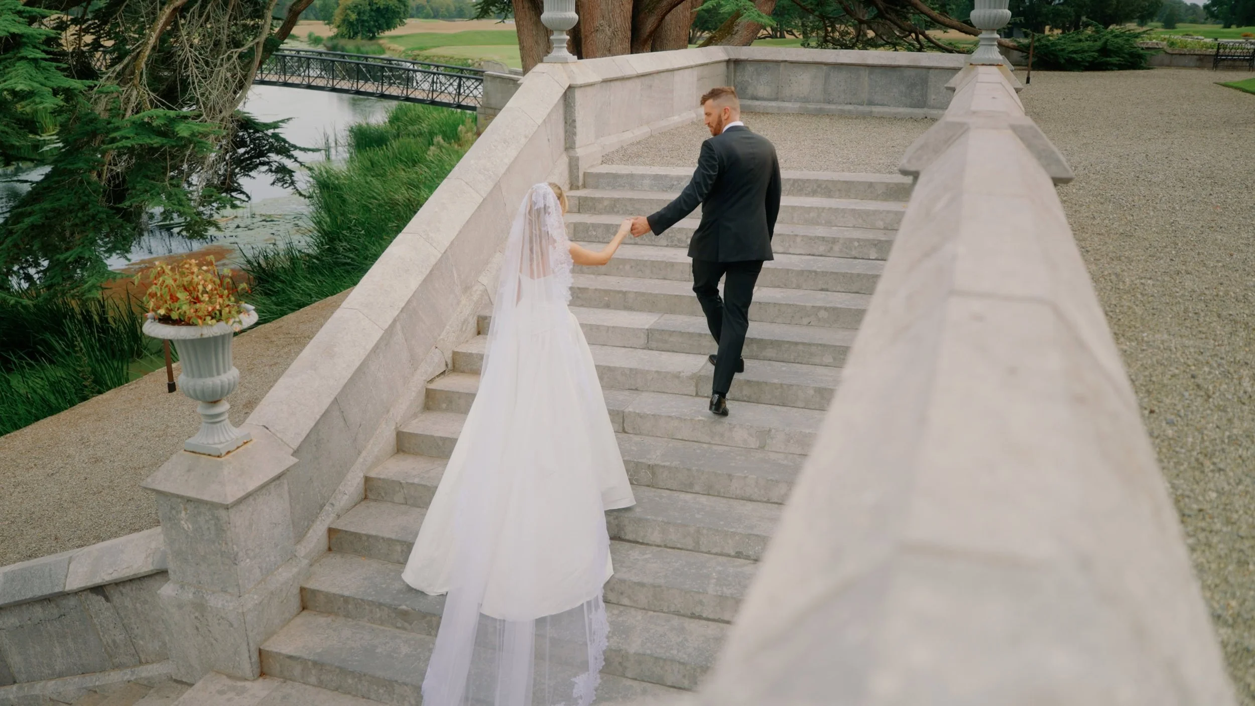 Bride and groom walking hand-in-hand up the stone terrace steps at Adare Manor, County Limerick, with the River Maigue and estate grounds in the background — captured by Dear Oliver wedding videographer
