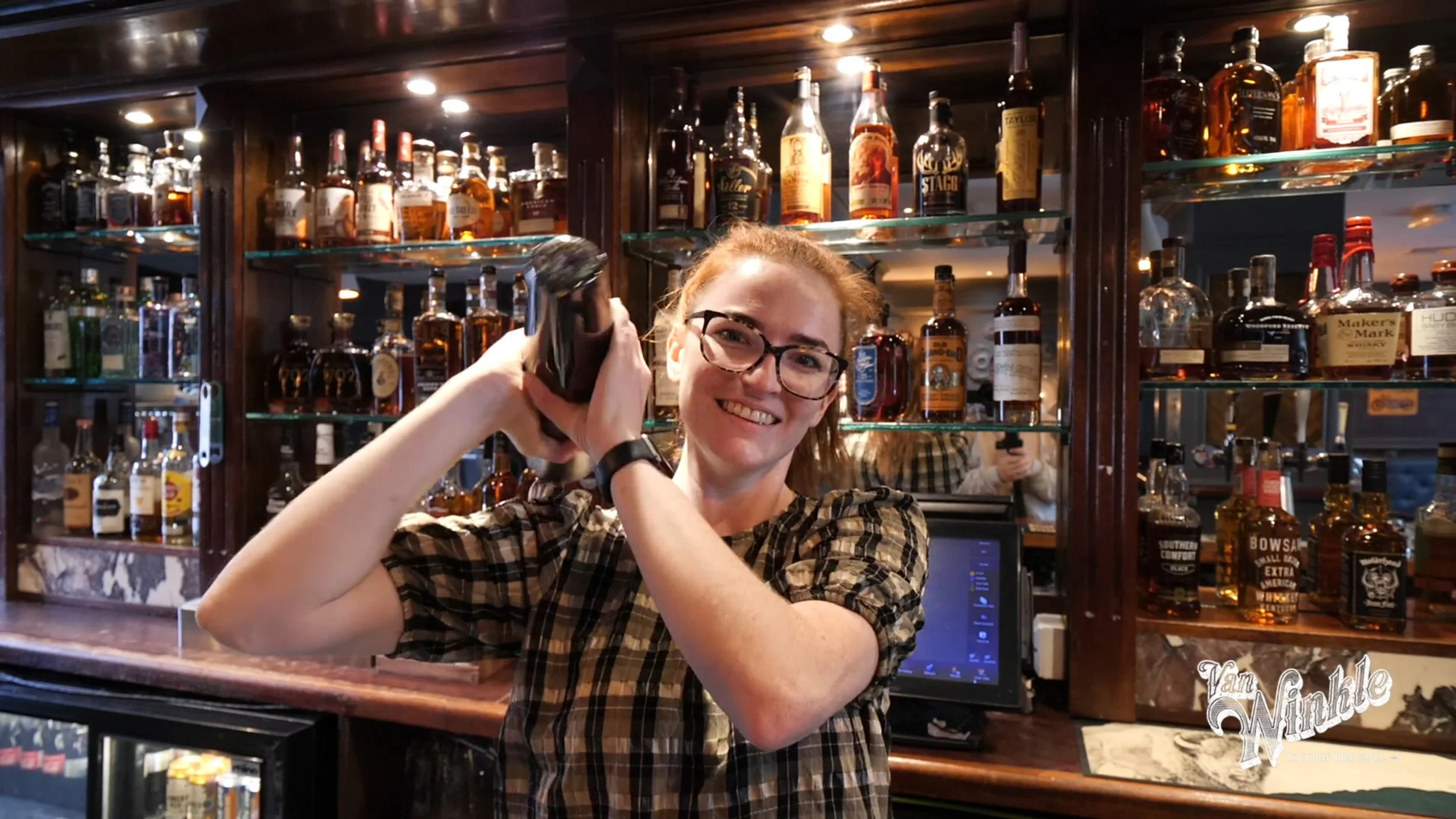 Bartender smiling and shaking a cocktail shaker behind a bar with various liquor bottles on display.