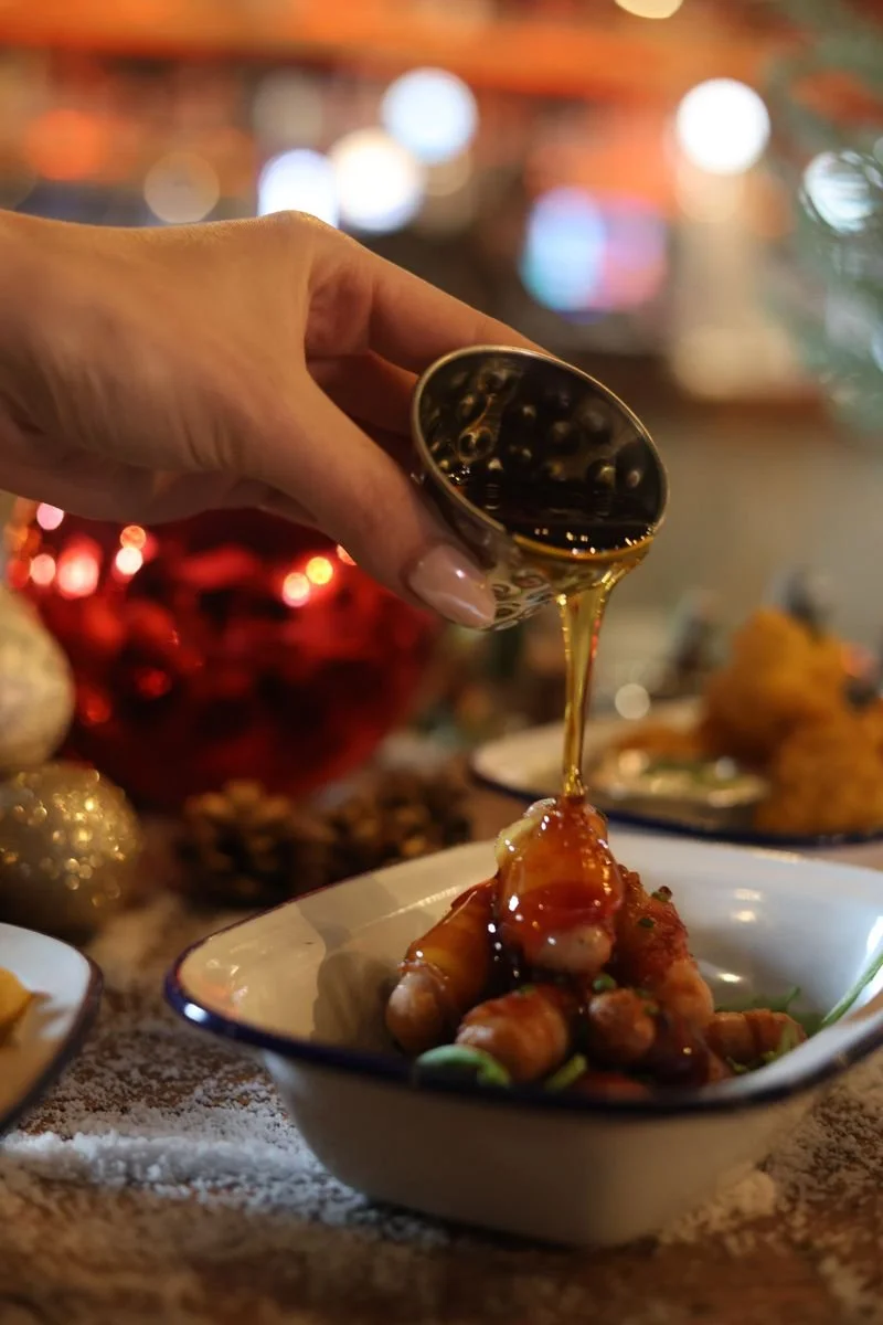 Syrup being poured over bacon-wrapped sausages in a bowl, with festive decorations in the background.