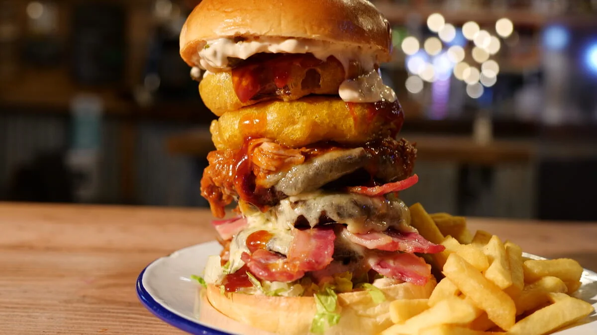 A large Van Winkle  burger with multiple beef patties, cheese, bacon, onion rings, and sauce, served with fries on a white plate.