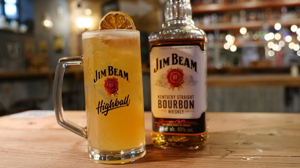 Jim Beam bourbon bottle and highball glass with drink and lemon slice on a bar table.