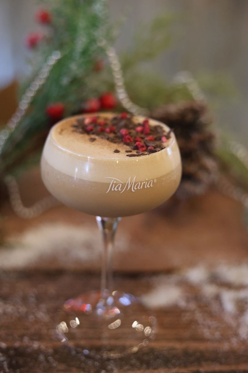 A frothy Tia Maria cocktail in a stemmed glass, garnished with chocolate flakes and red berries, with festive greenery in the background.