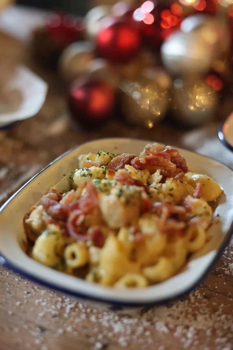 Baked macaroni and cheese with bacon in an enamel dish, decorated with herbs, on a wooden table with Christmas ornaments in the background.