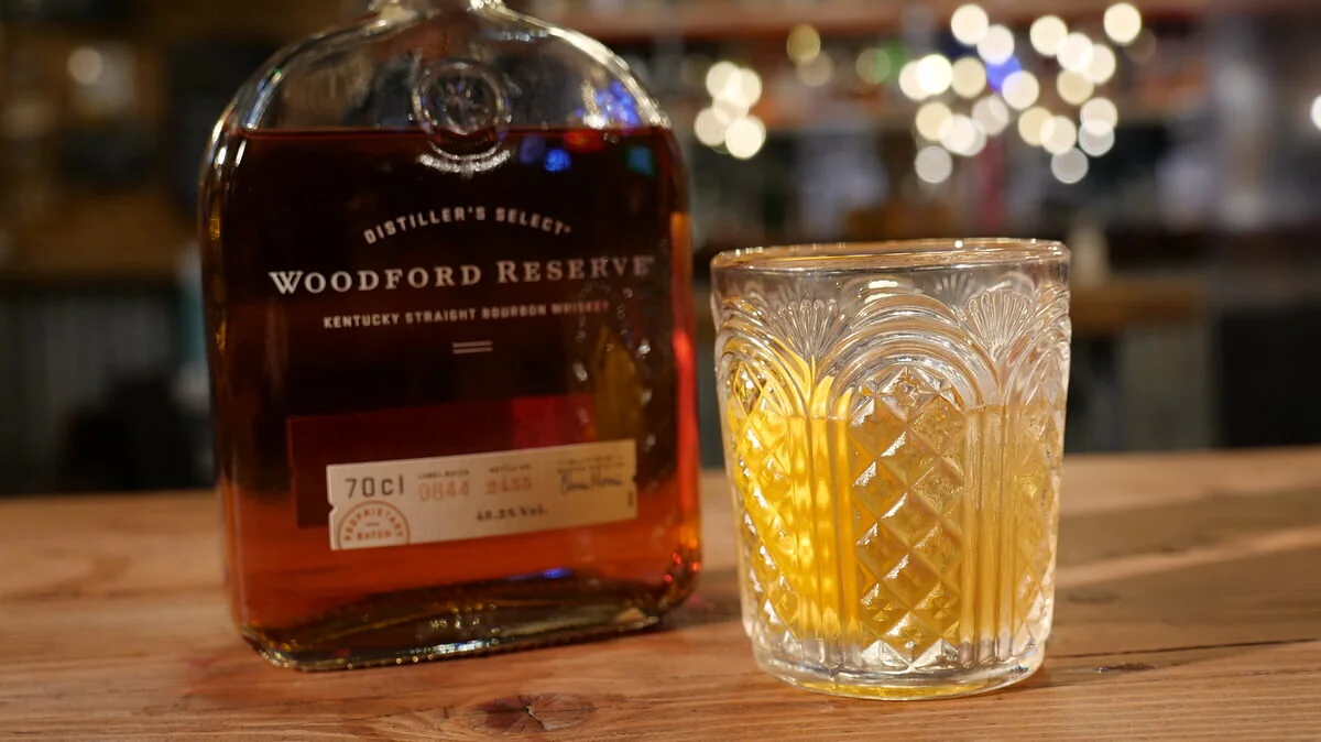 Bottle of Woodford Reserve bourbon and a glass filled with bourbon on a wooden table with a blurred background.
