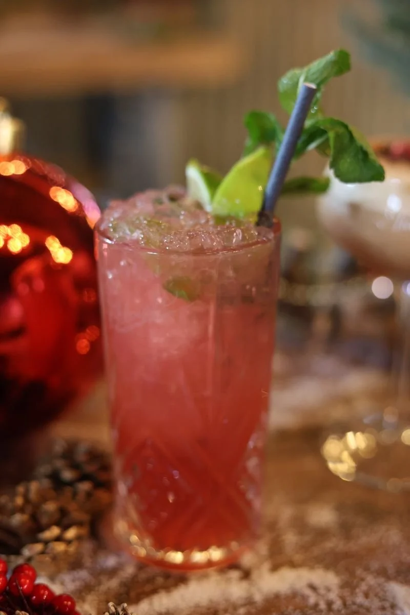 Pink cocktail with lime and mint garnish in a glass, surrounded by festive decor.