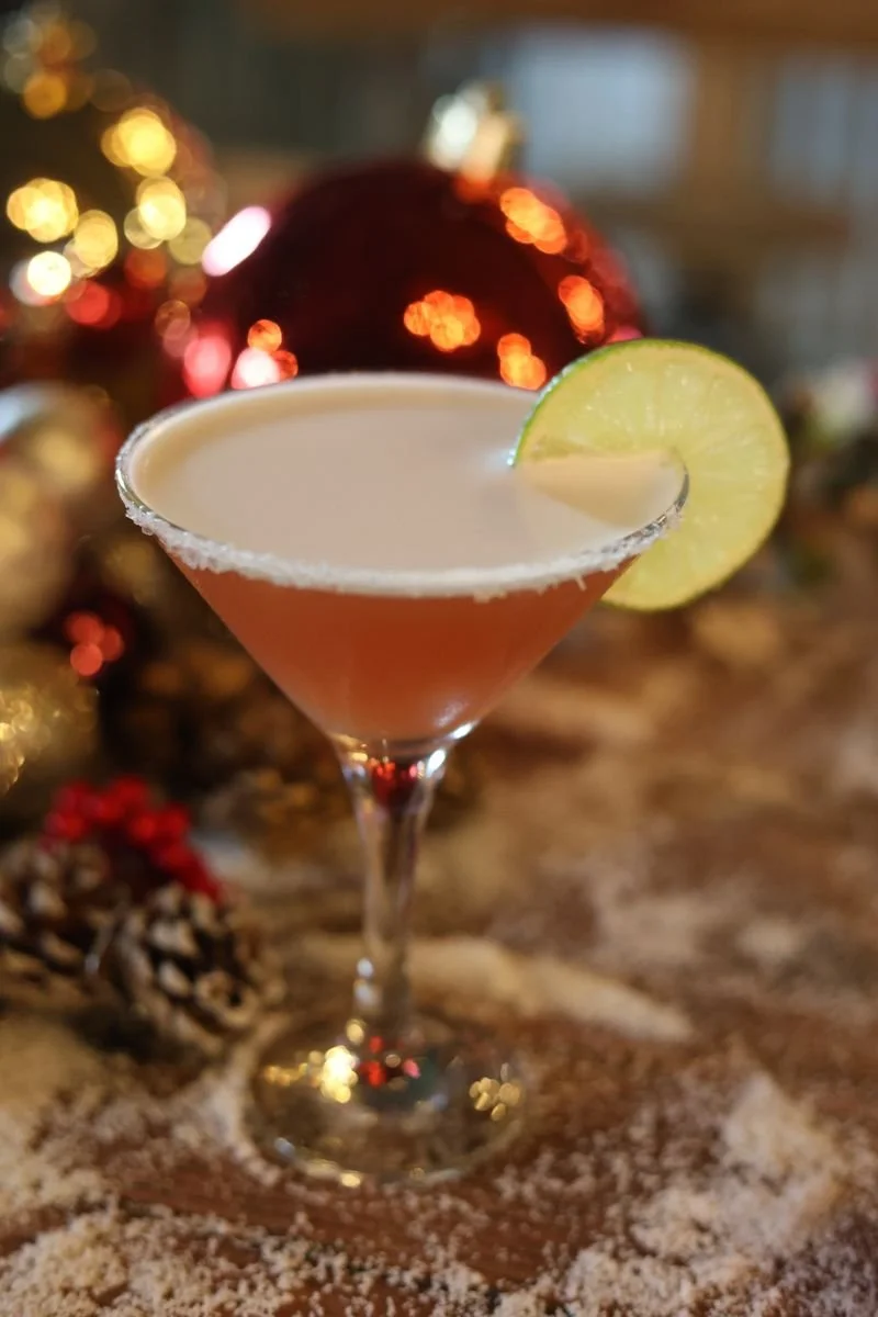 Holiday cocktail in a martini glass with lime garnish and sugar rim, surrounded by festive decorations.