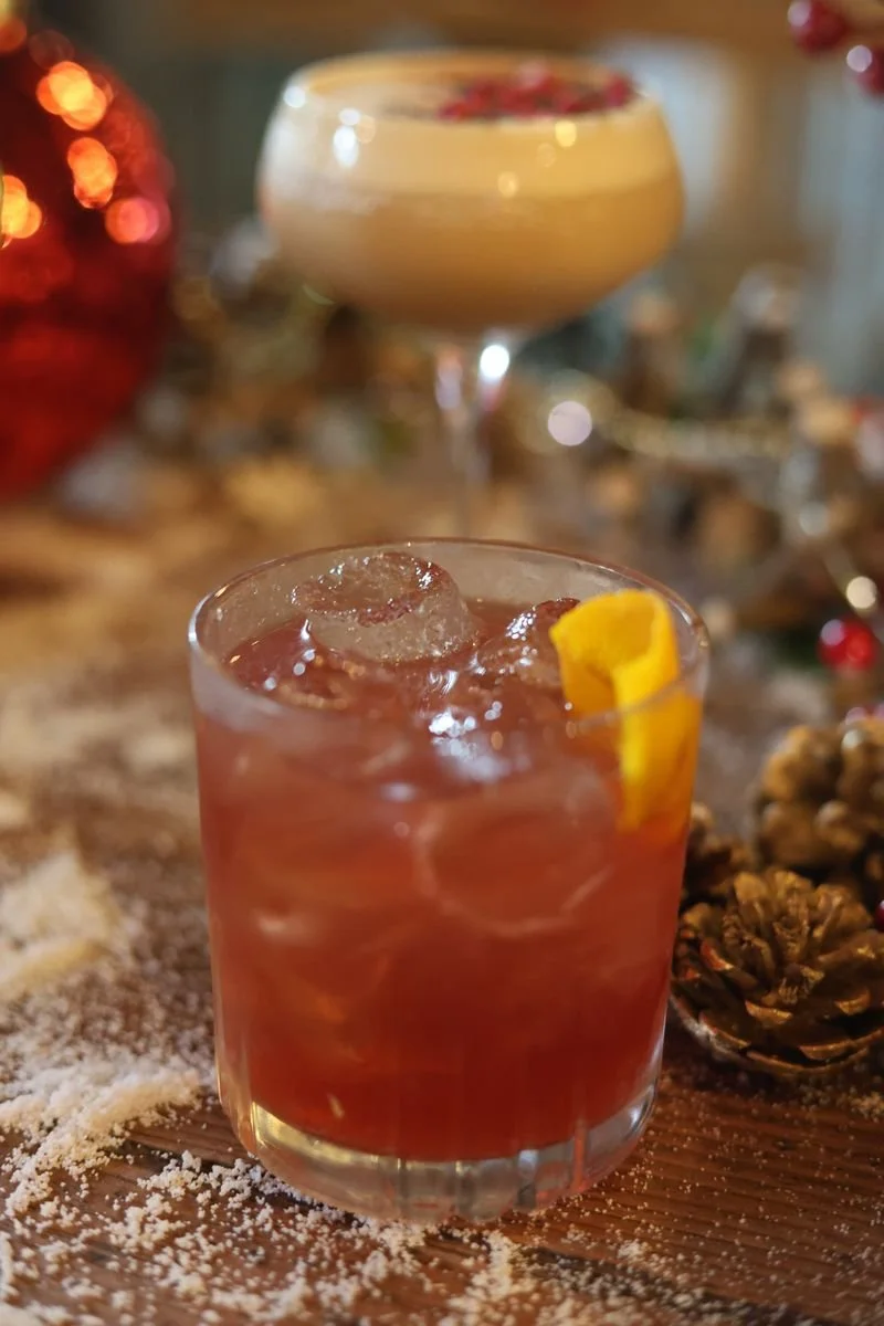 Close-up of a cocktail with ice and orange peel in a glass, with another creamy drink in the background, surrounded by festive decorations and pine cones.