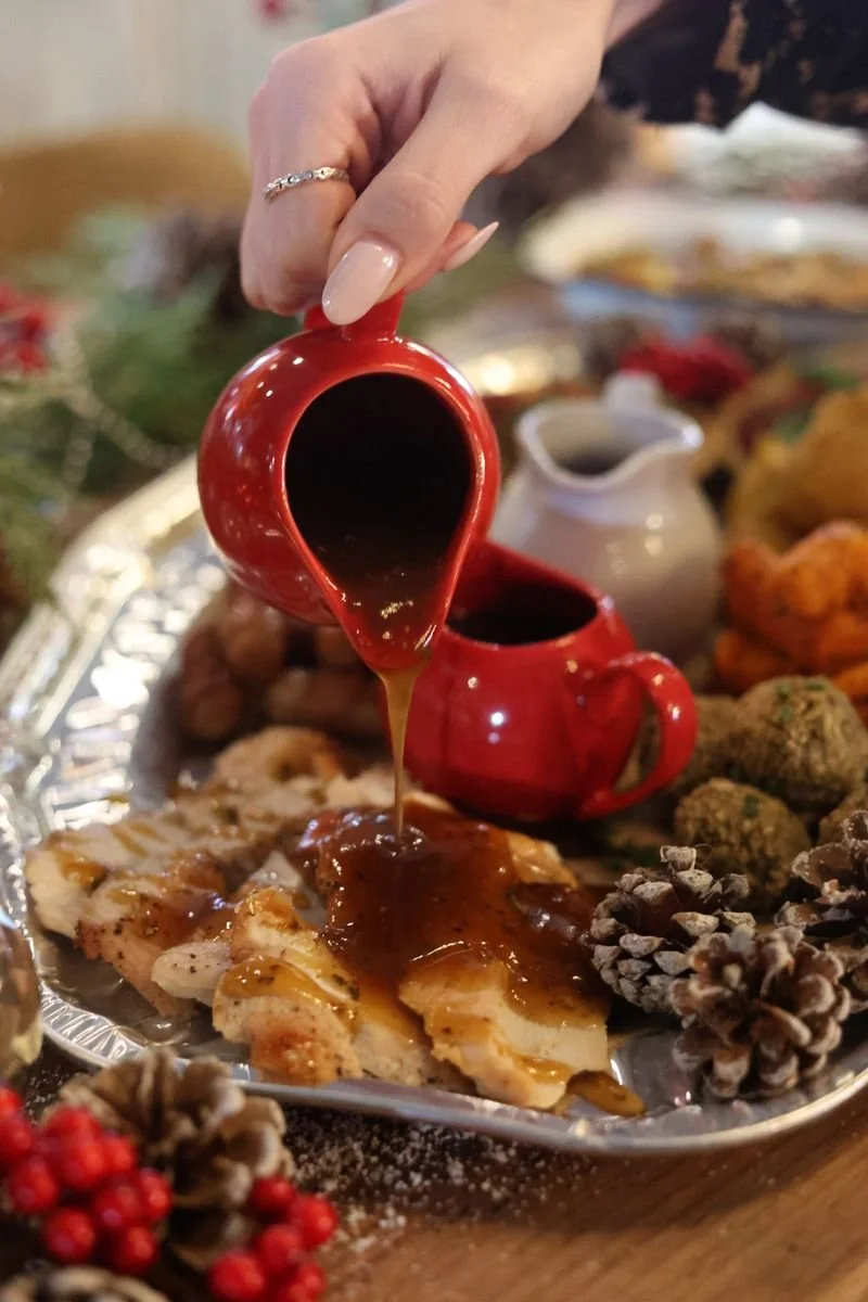 A hand pouring gravy from a red pitcher onto sliced turkey on a platter, surrounded by holiday decorations including pinecones and berries.
